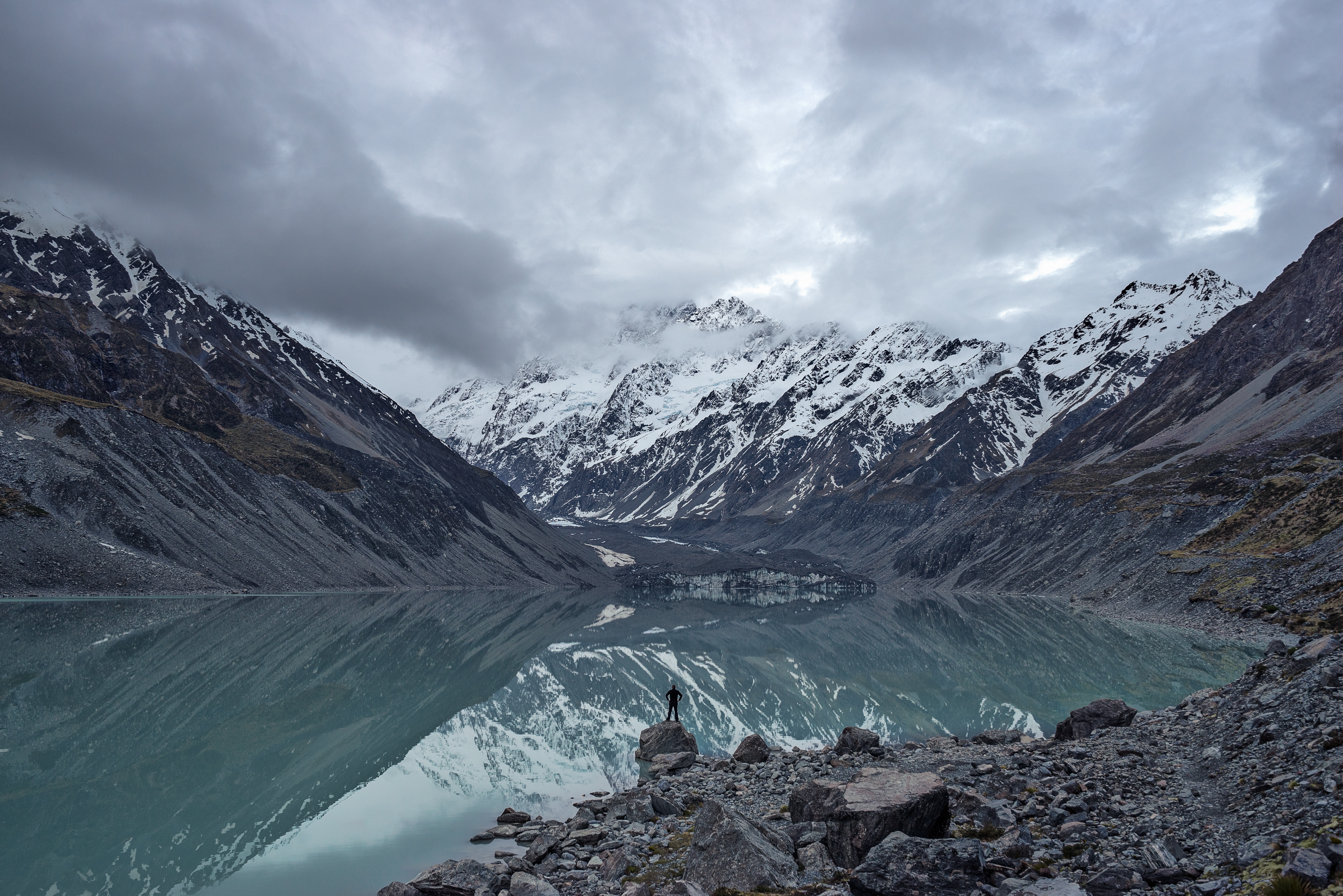 Person standing on rocks by a lake with snow-capped mountains HD Wallpaper