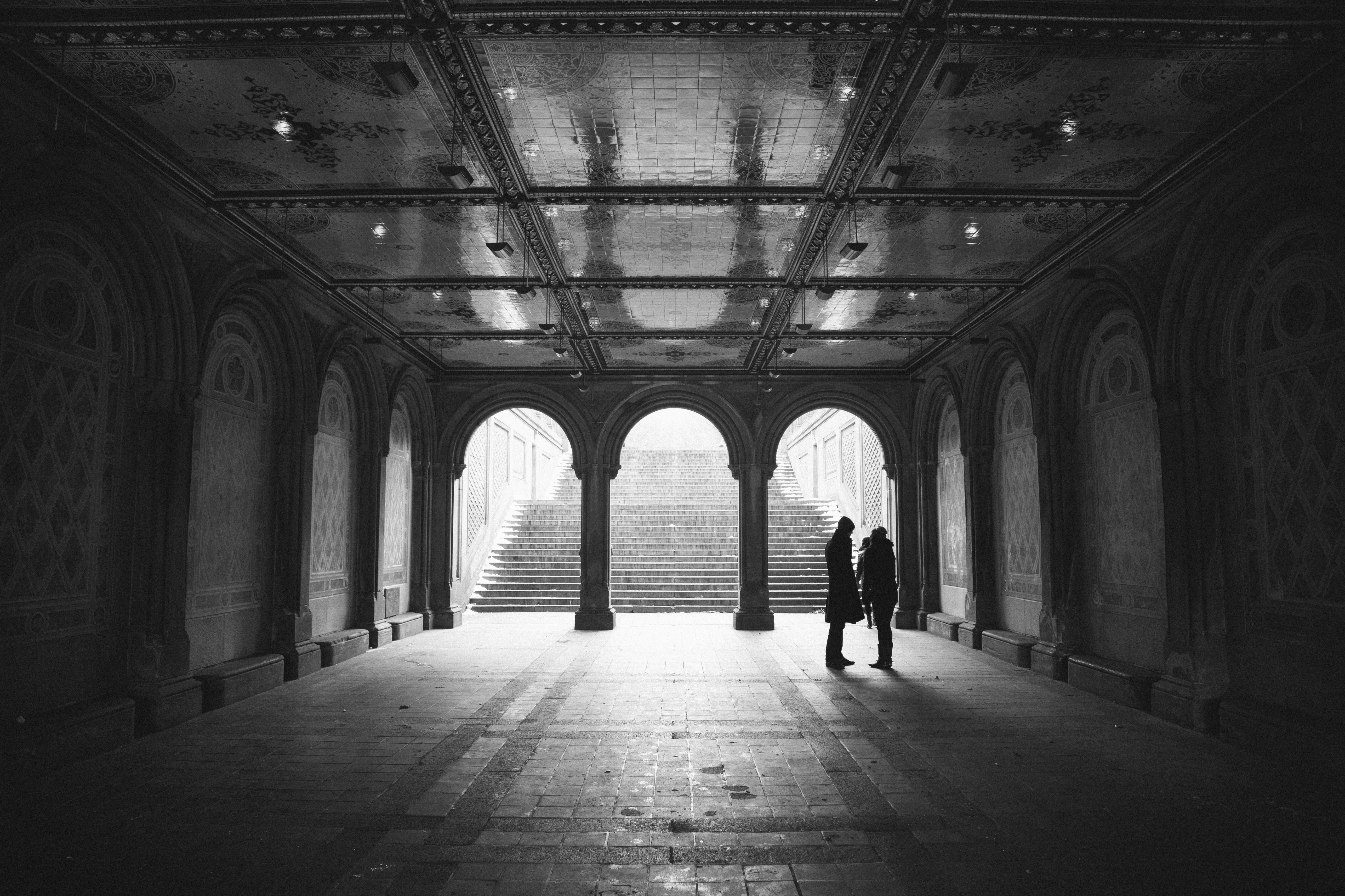 Two people stand in a grand arched hallway with stairs HD Wallpaper