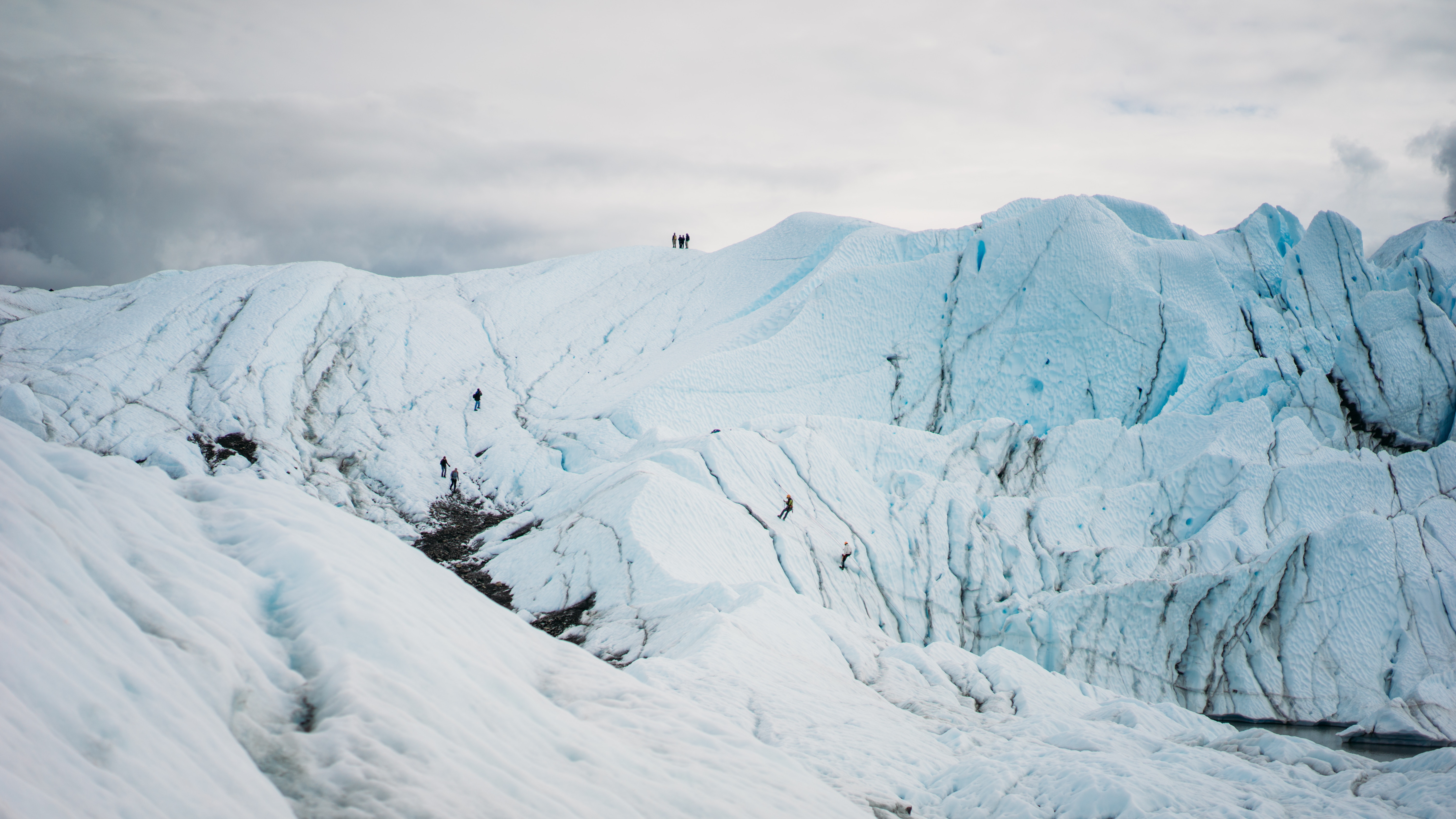 People climbing on a vast blue glacier under a cloudy sky HD Wallpaper