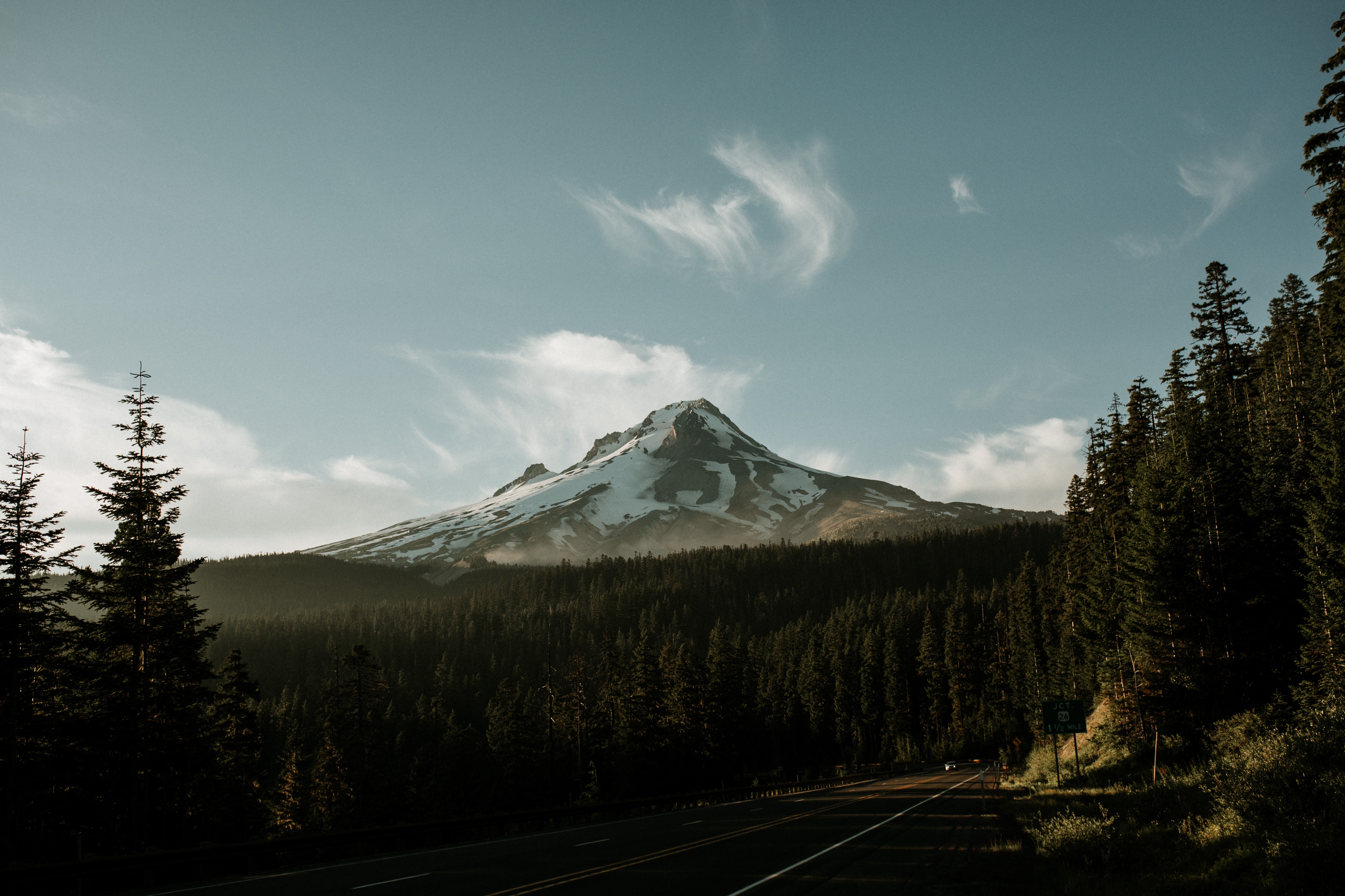 Snow-capped mountain peak above a dark forest and road HD Wallpaper