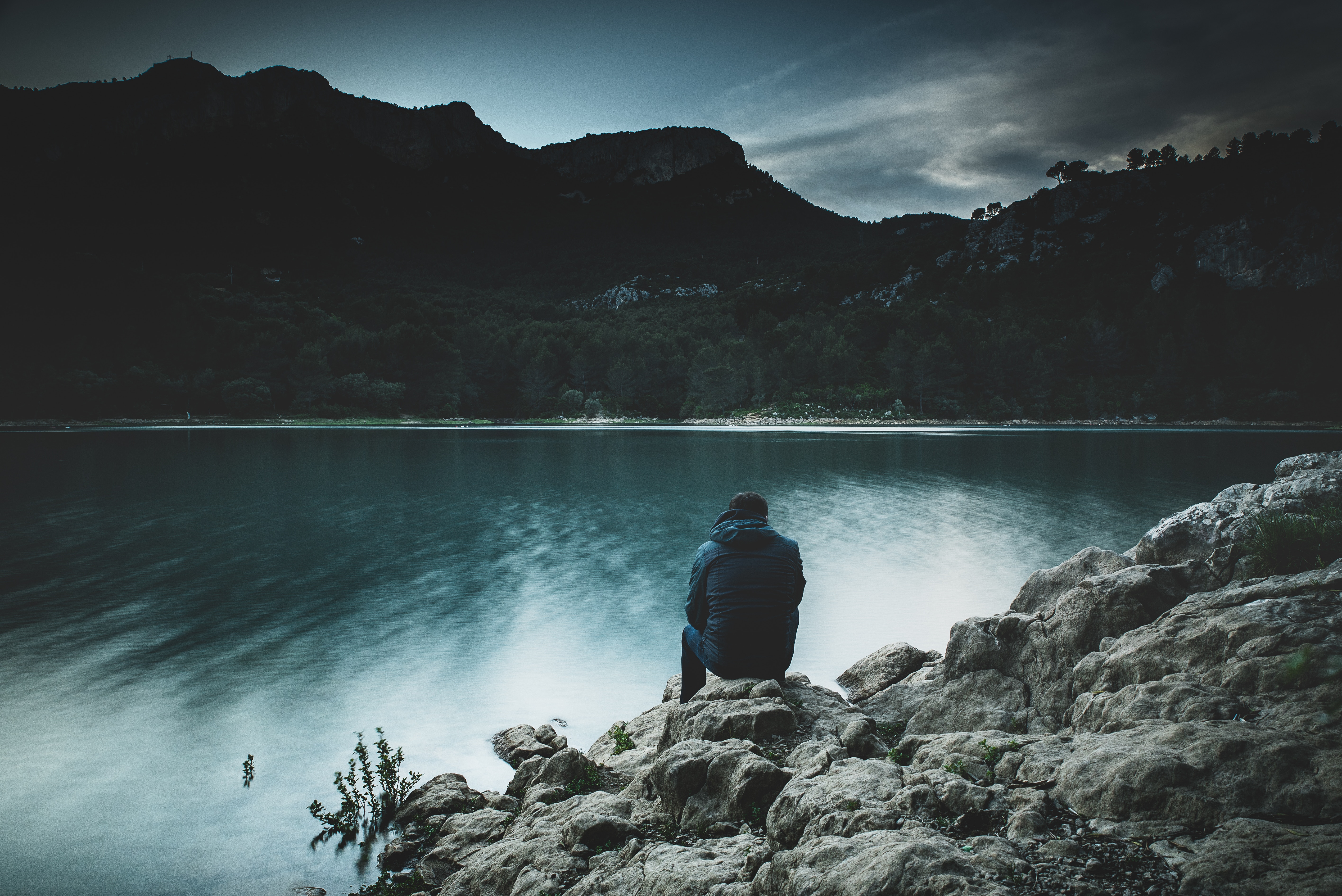 Man sitting on rocks by a lake with mountains HD Wallpaper