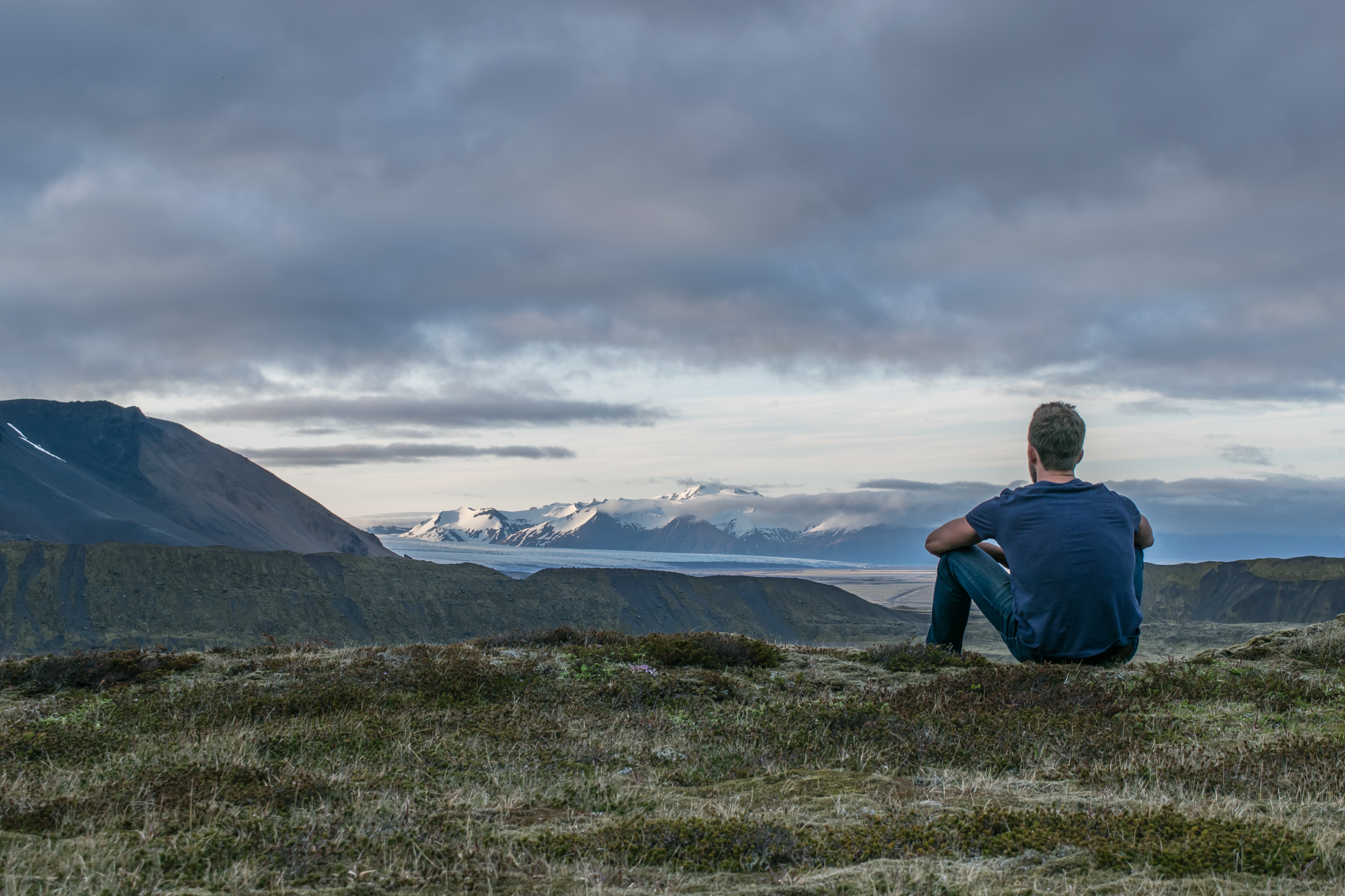 Man sitting on a grassy hill overlooking Icelandic mountains HD Wallpaper
