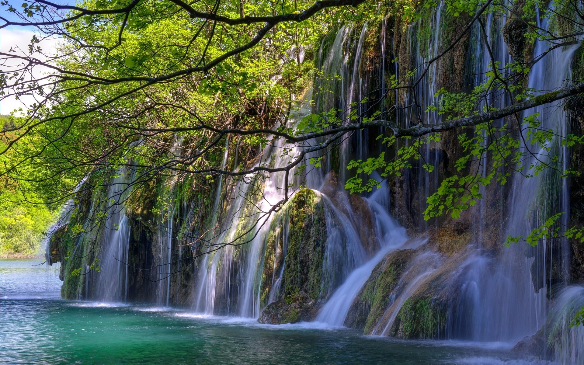 Waterfall cascading into a turquoise lake surrounded by greenery HD Wallpaper