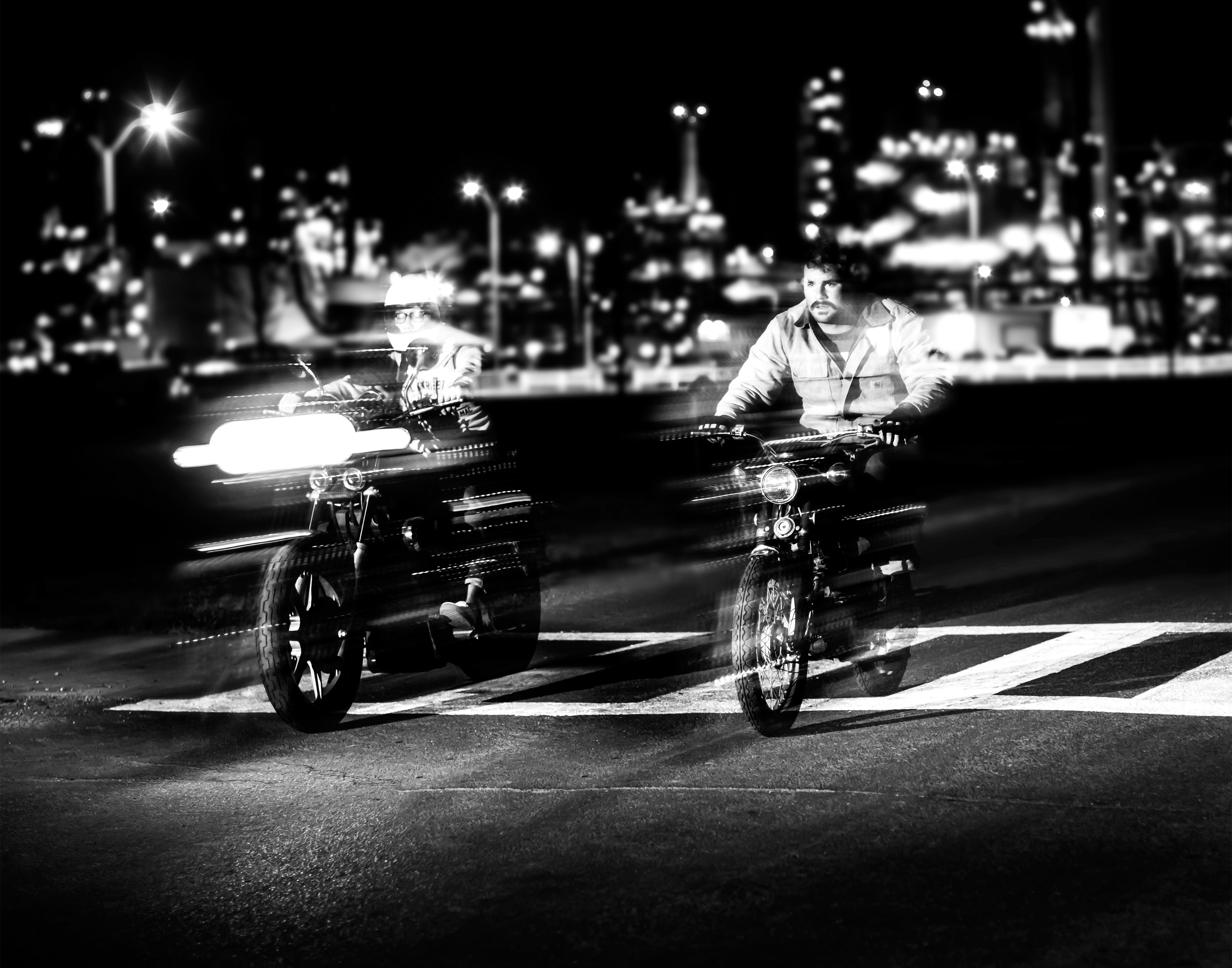 Two motorcyclists riding at night with city lights in background HD Wallpaper