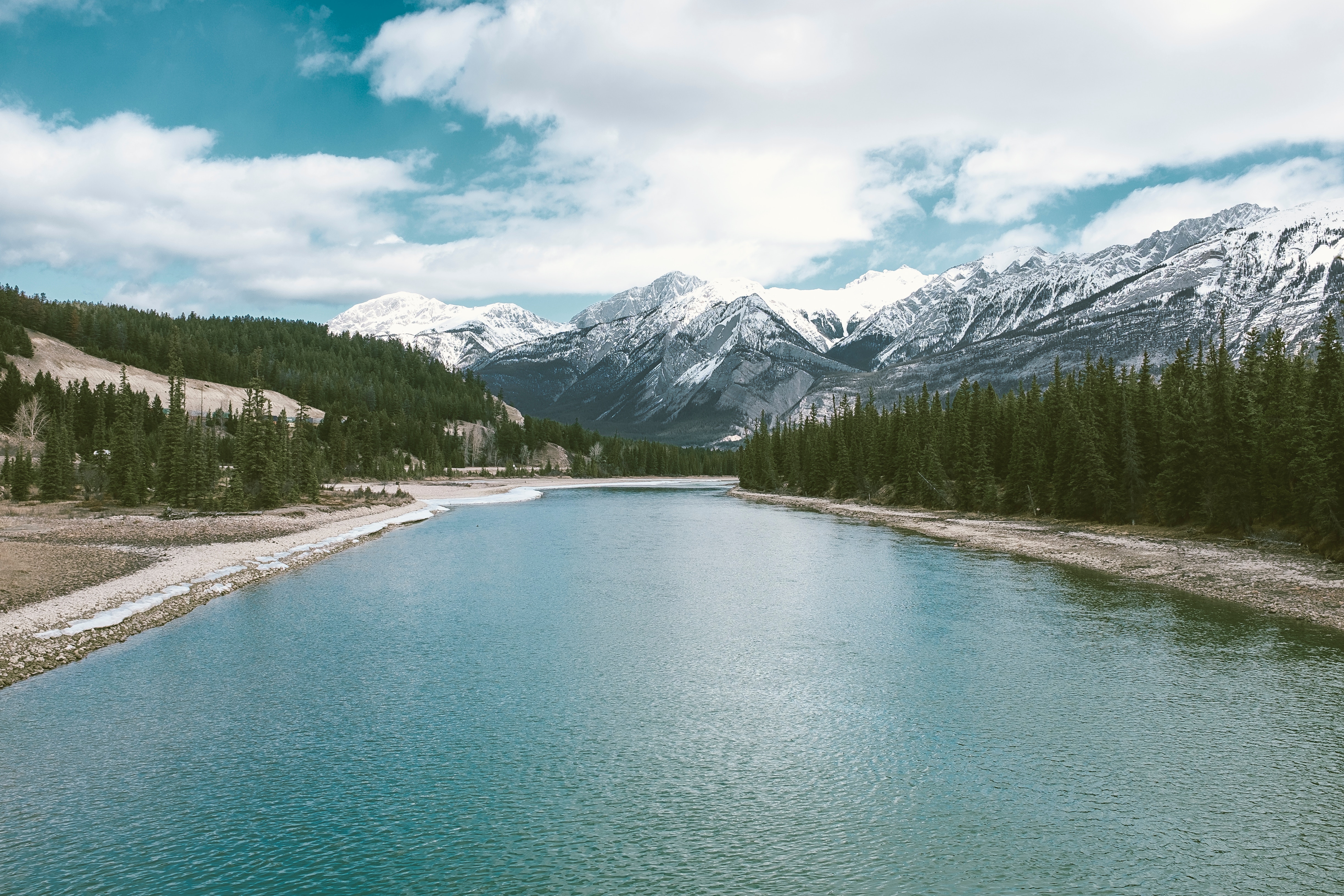 Turquoise river flowing through a forest with snow-capped mountains HD Wallpaper