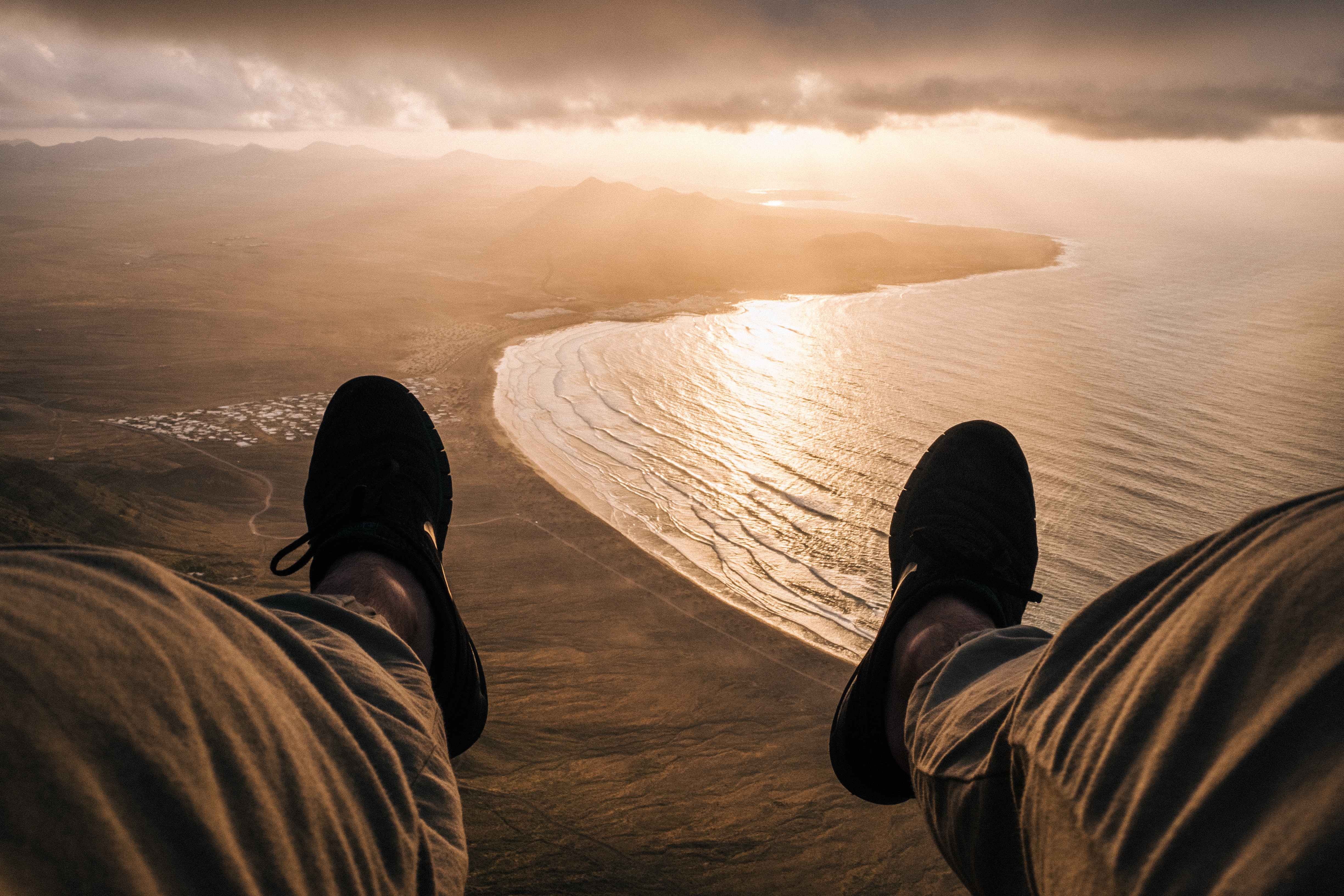 Person's legs dangling over a coastal landscape at sunset HD Wallpaper