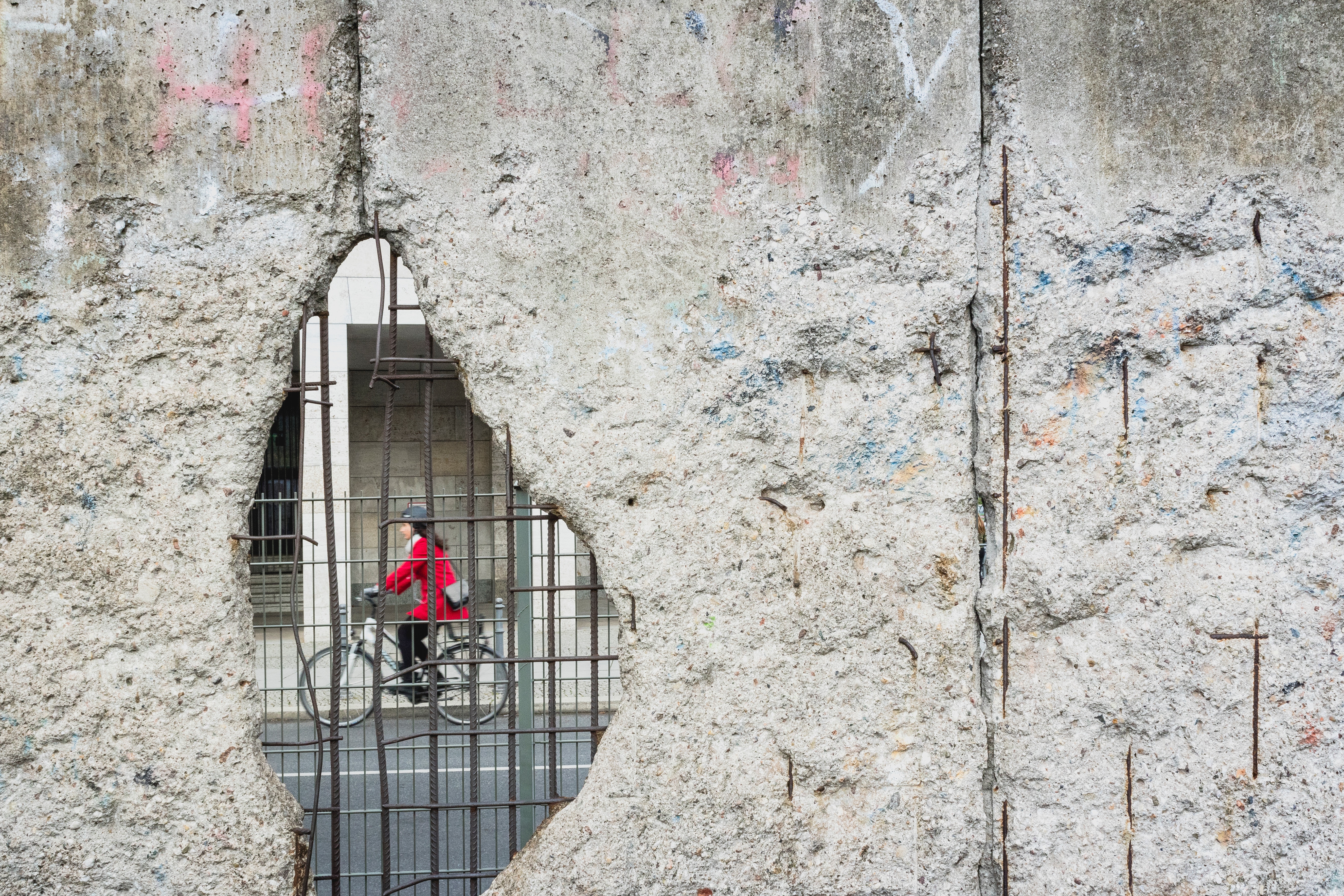 Woman in red coat cycling through a hole in the Berlin Wall HD Wallpaper