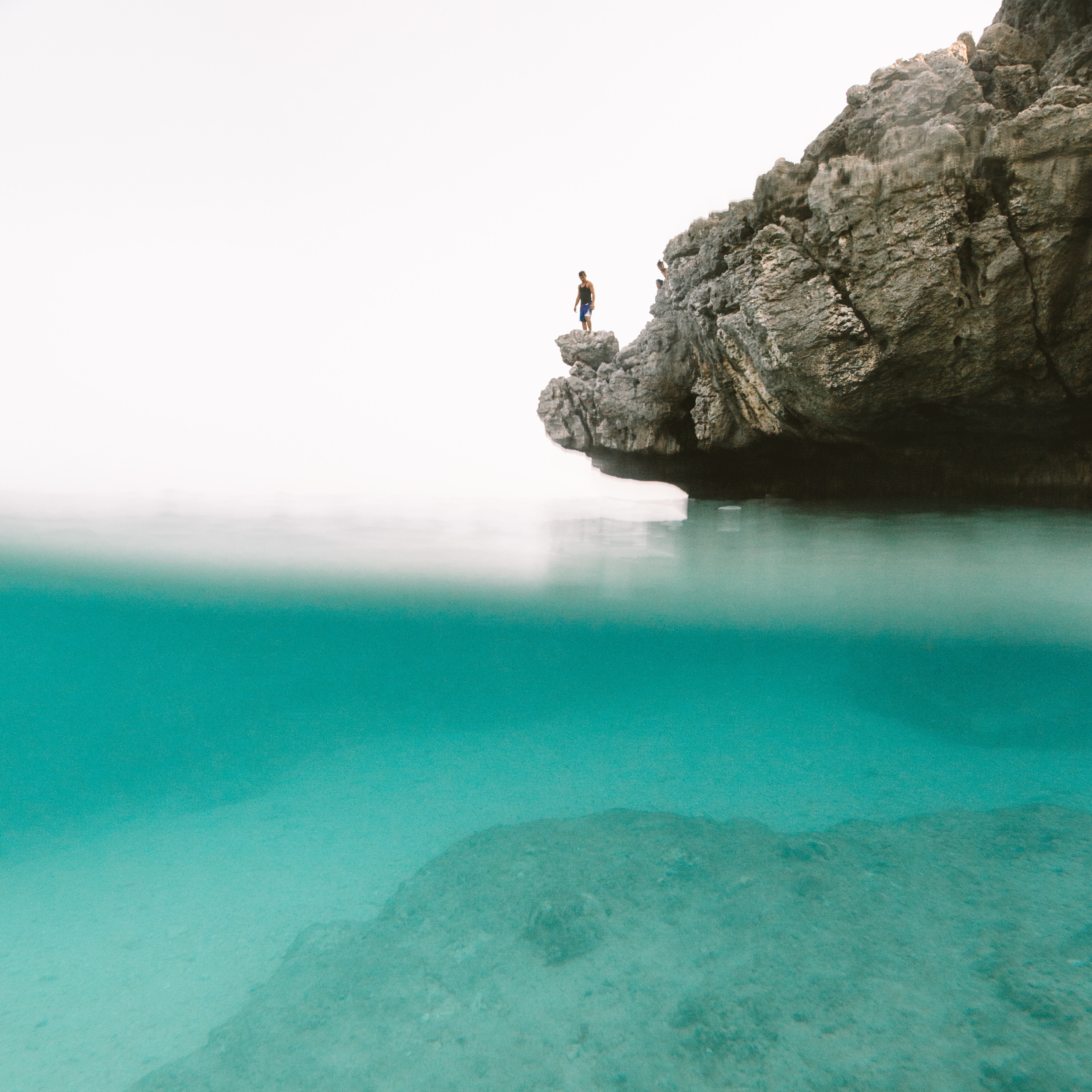 Person standing on a rock cliff above turquoise water HD Wallpaper