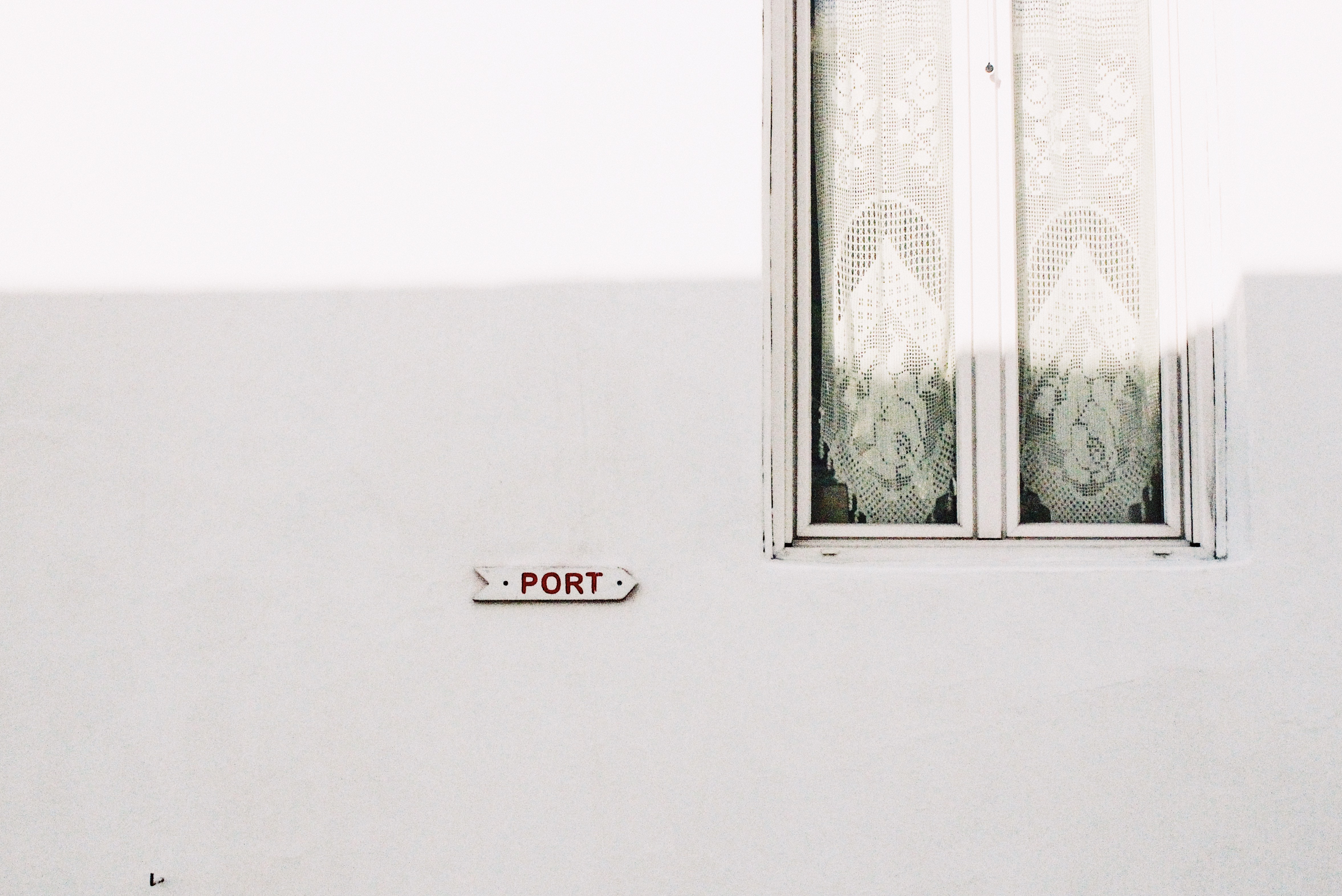 White wall with a sign pointing to the port and a window with lace curtains HD Wallpaper