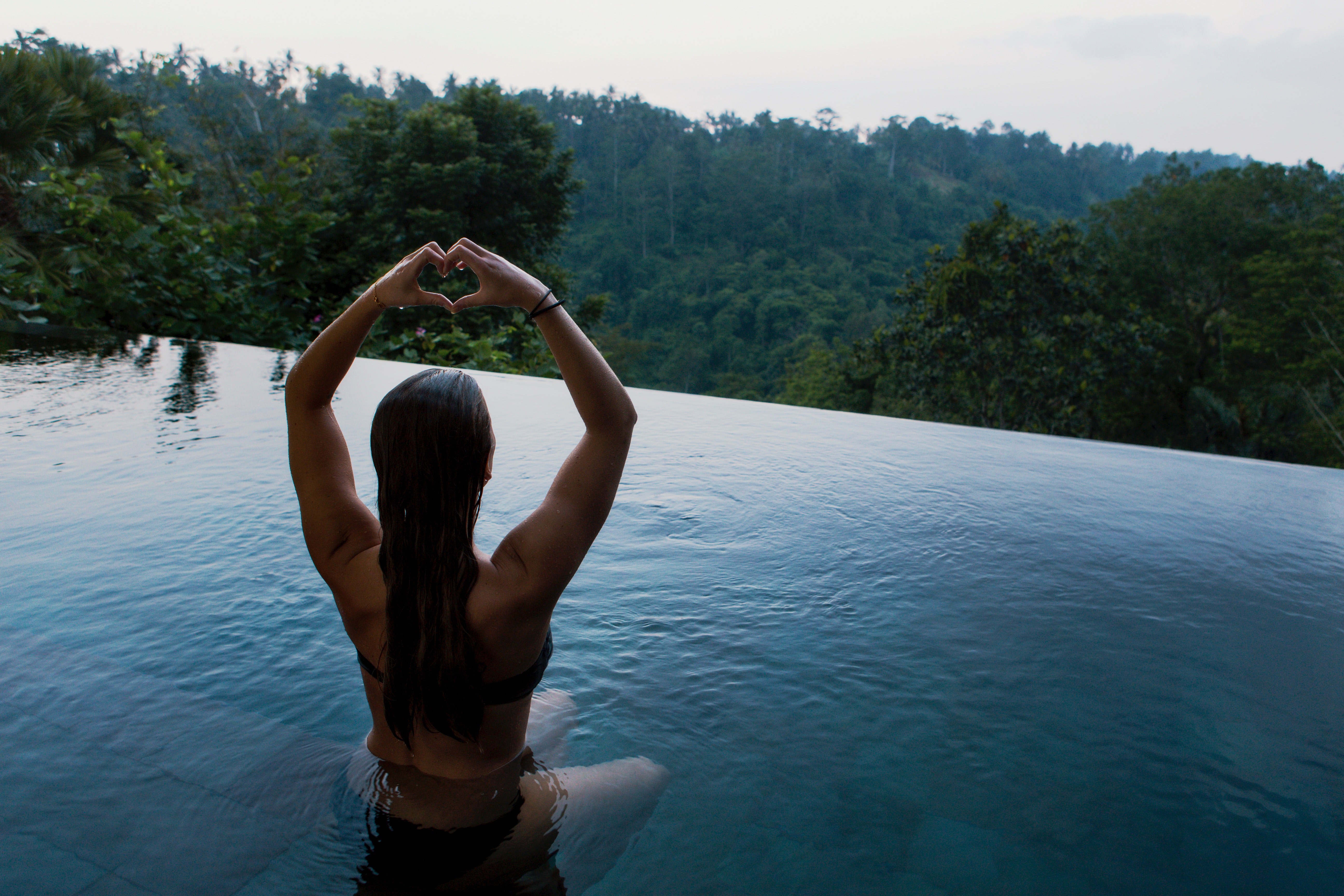 Woman making heart shape in infinity pool overlooking jungle HD Wallpaper