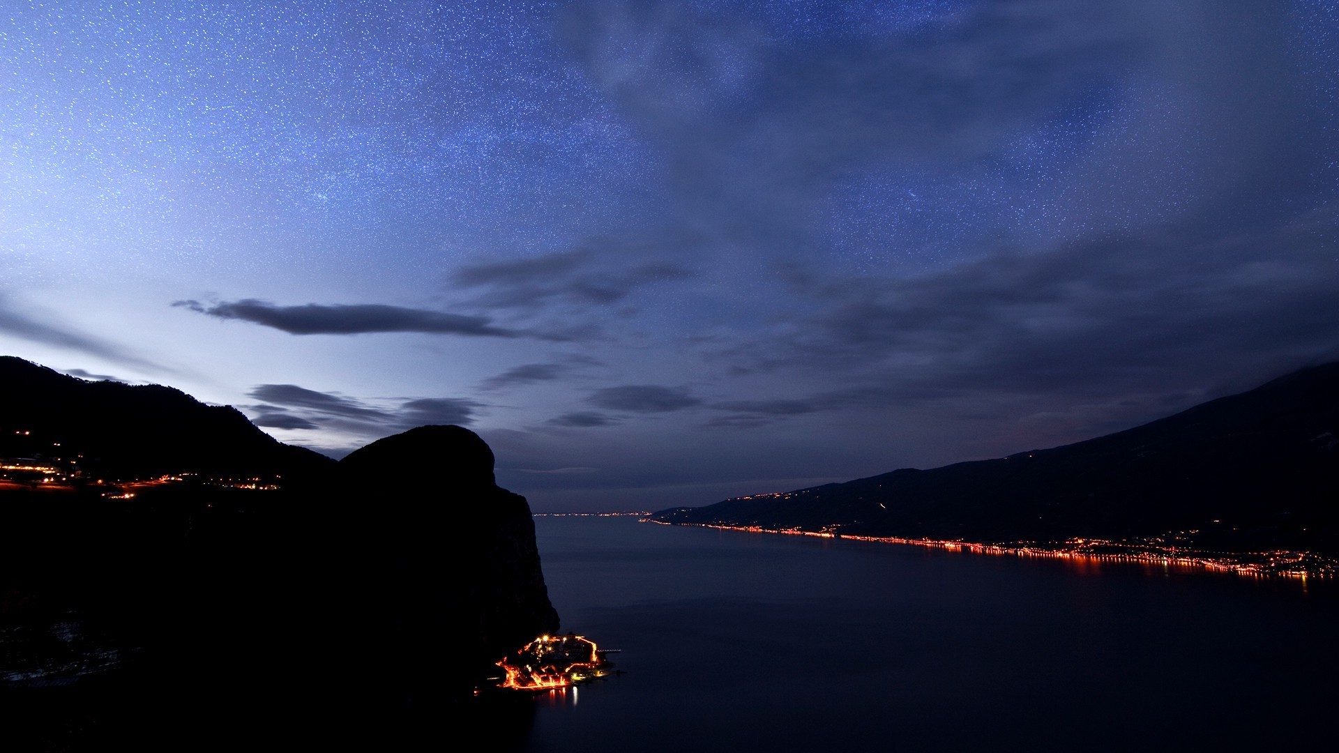 Night sky over a lake with illuminated towns HD Wallpaper