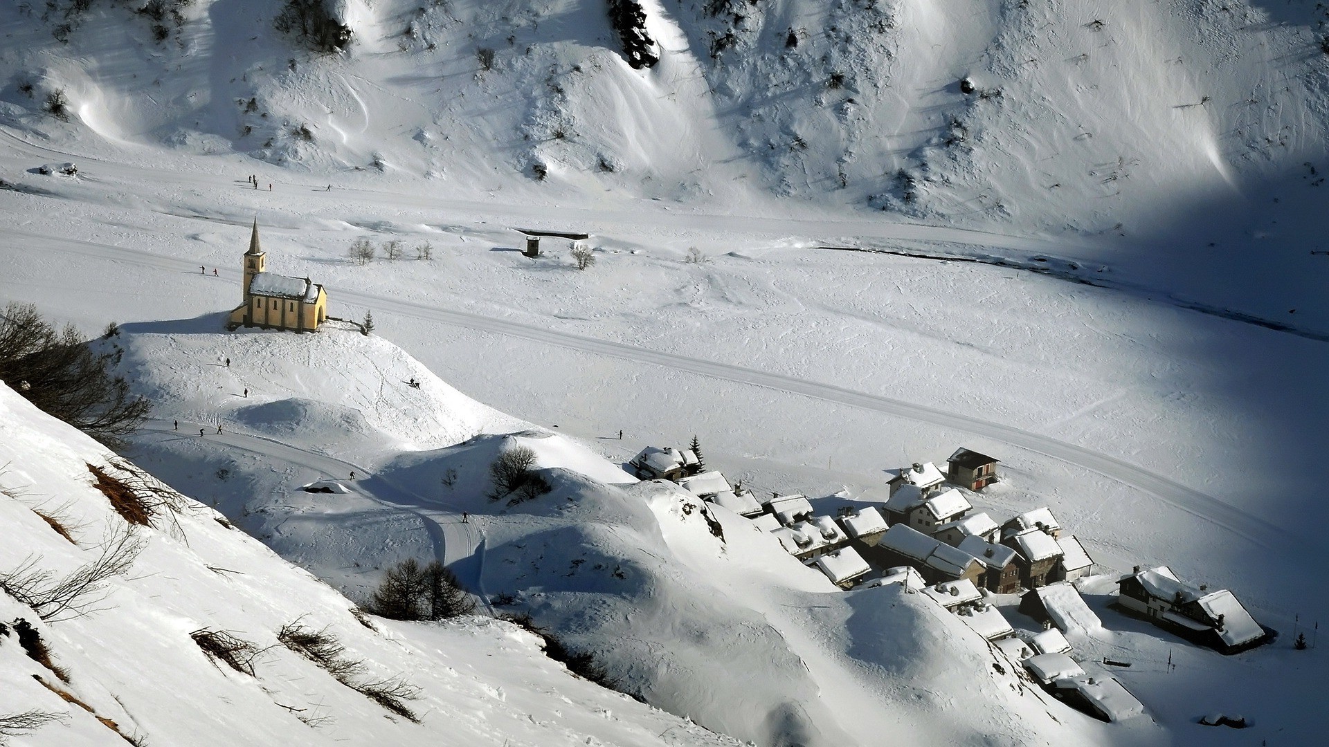 Snowy mountain village with church and skiers HD Wallpaper