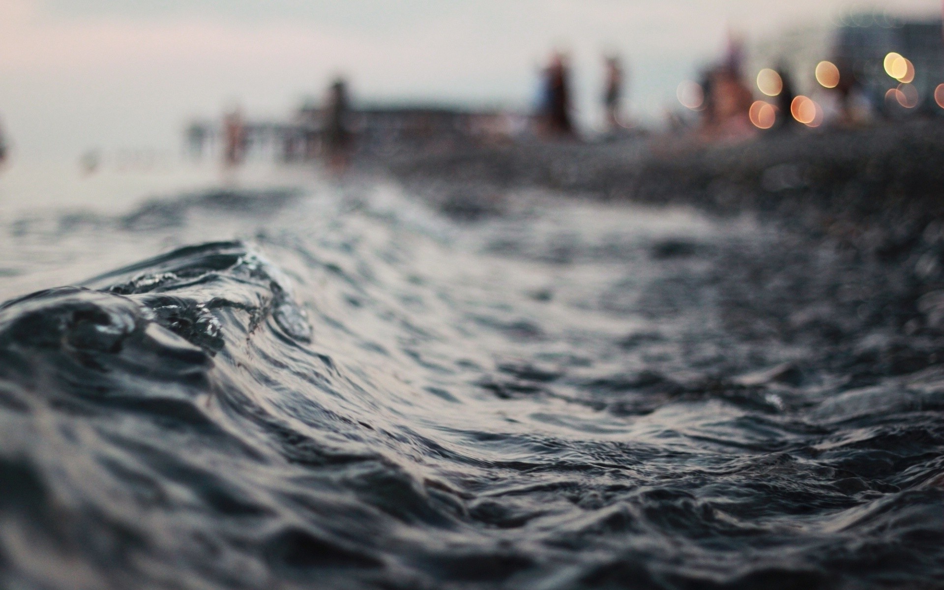 Close-up of a wave with blurred beachgoers in the background HD Wallpaper