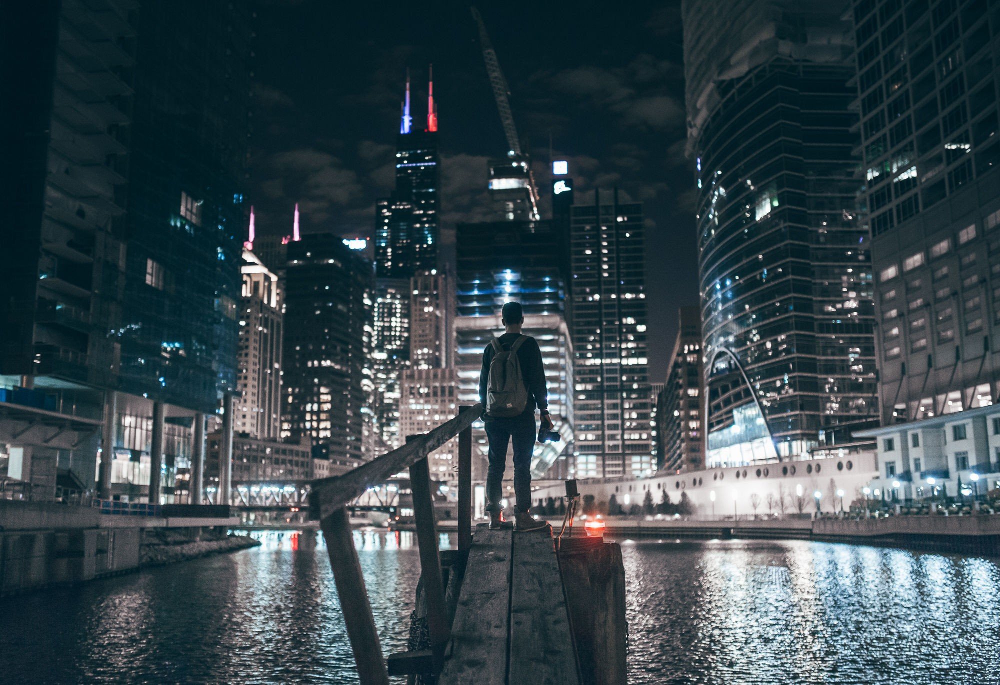 Man with backpack and camera on wooden pier overlooking Chicago cityscape at night HD Wallpaper