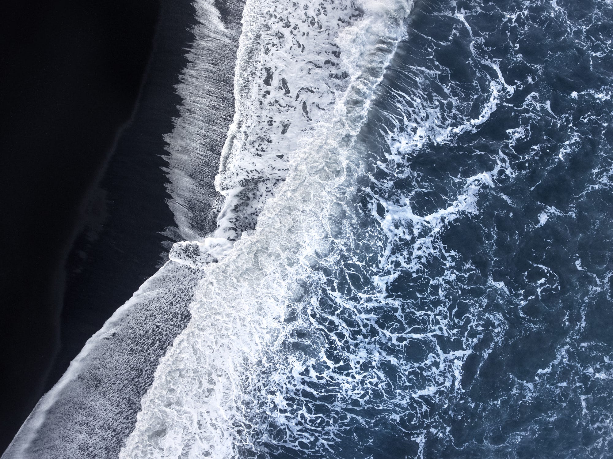 Aerial view of waves crashing on a black sand beach HD Wallpaper