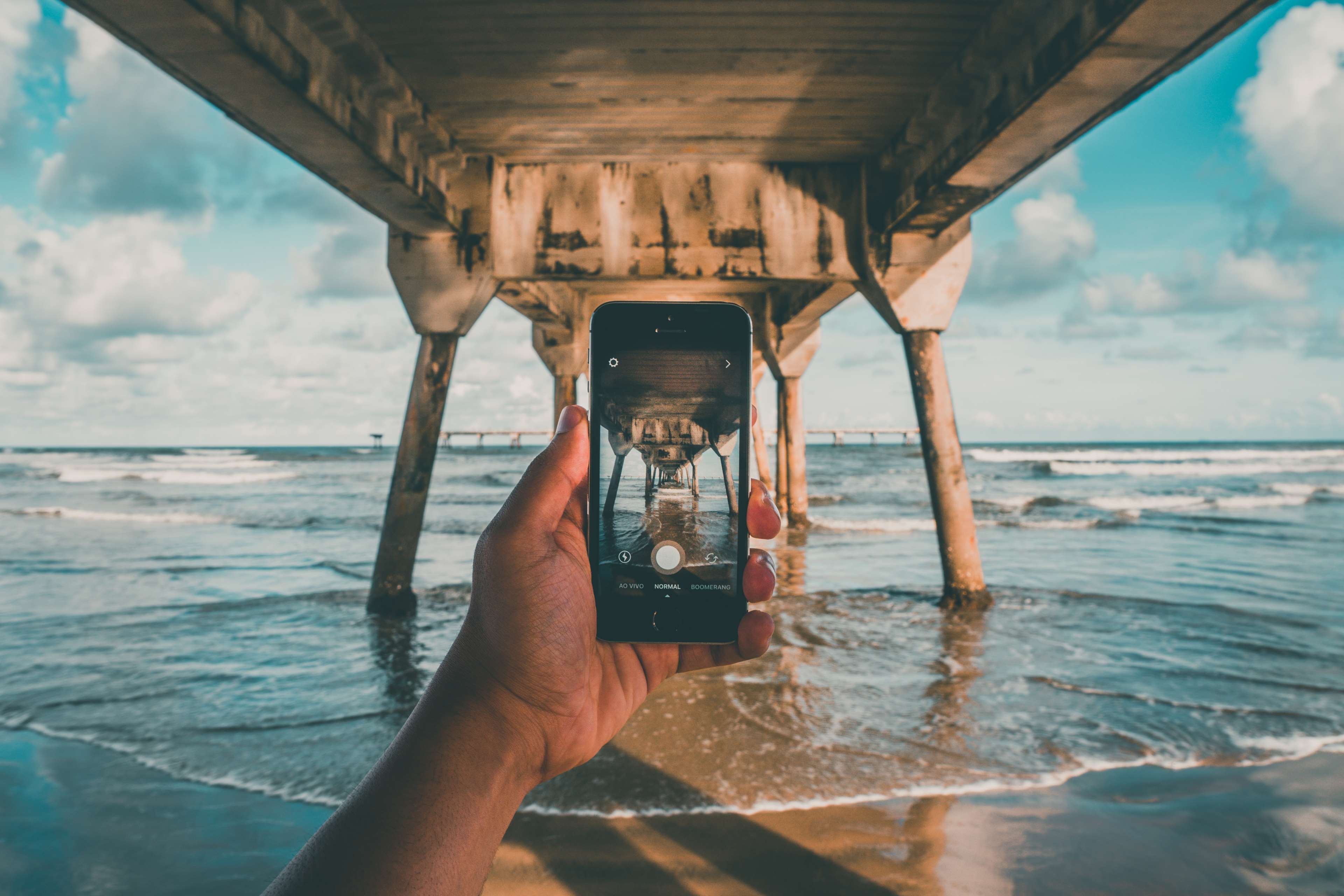 Hand holding phone taking picture of pier over ocean HD Wallpaper