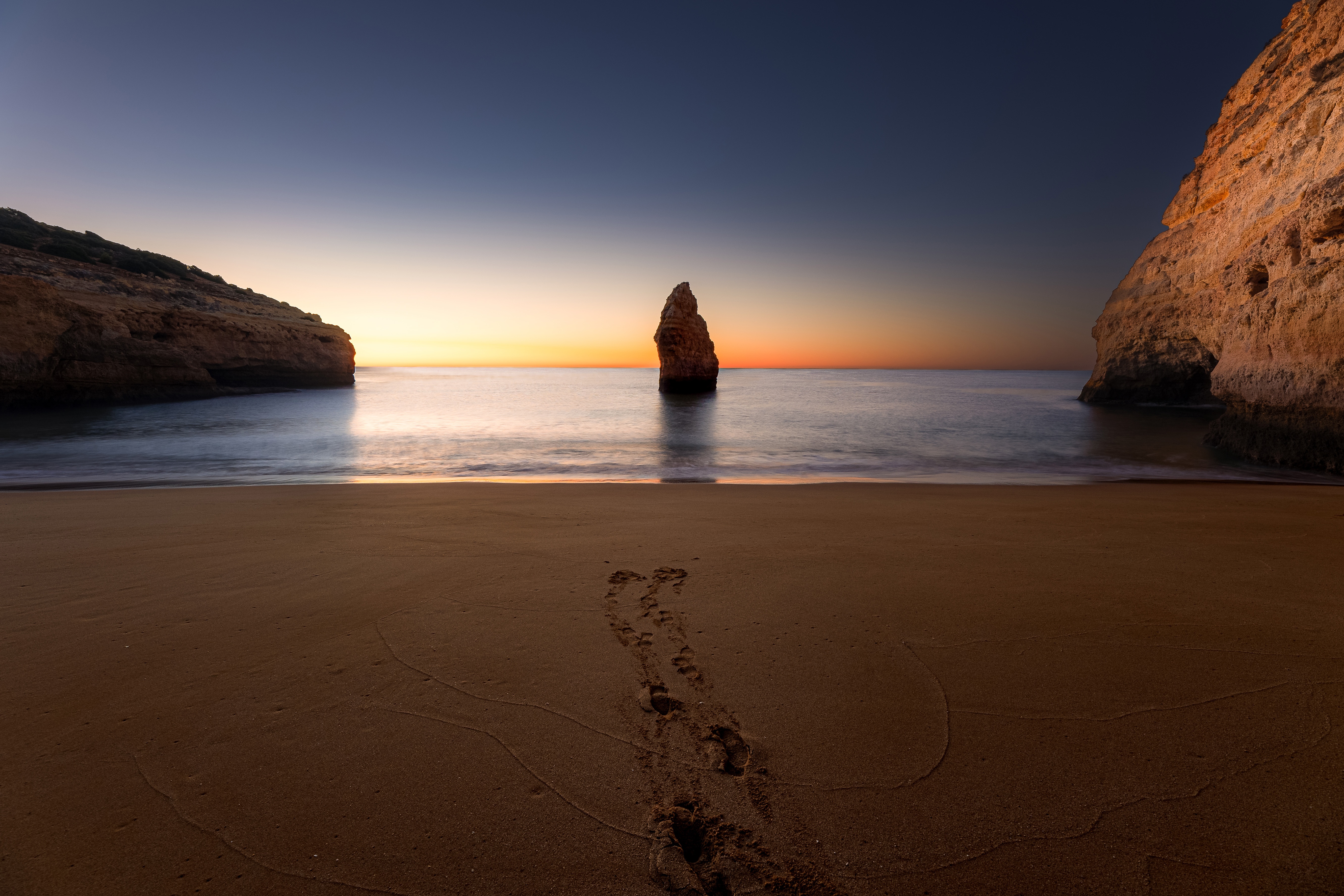 Beach sunset with rock formation and footprints HD Wallpaper