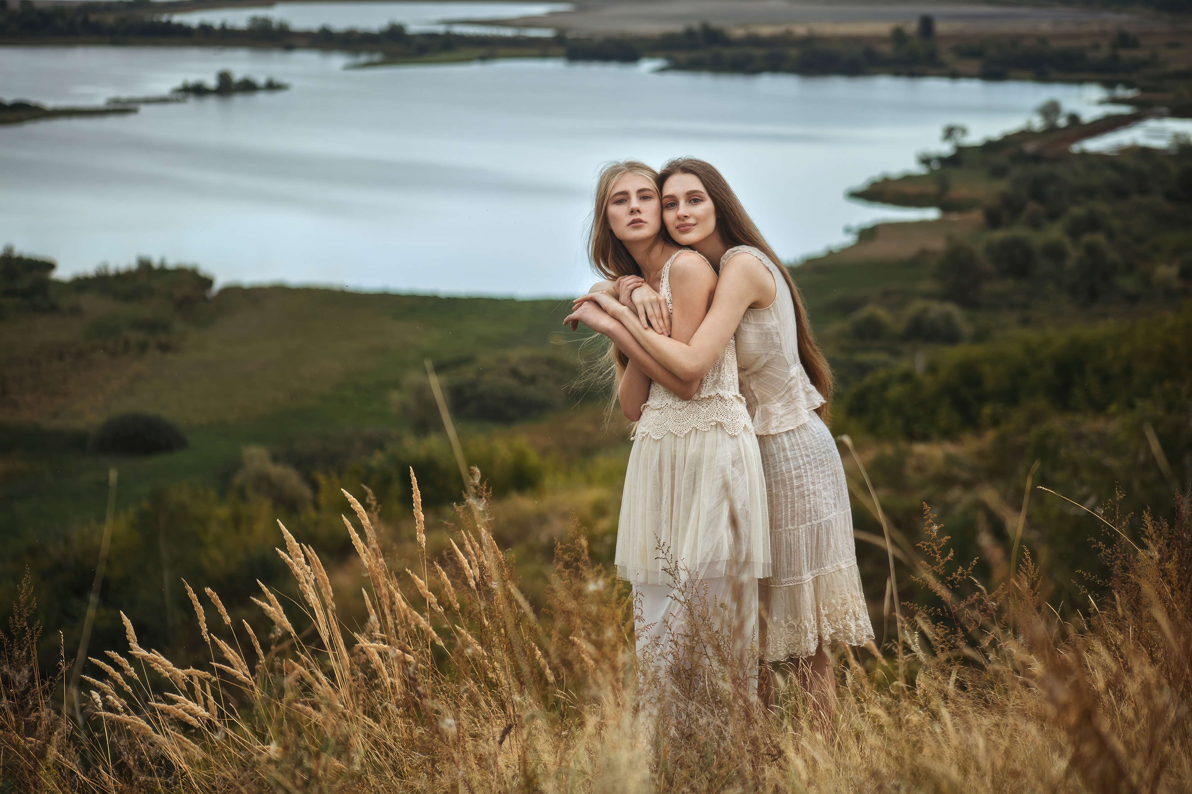 Two women embracing in a field with a lake HD Wallpaper