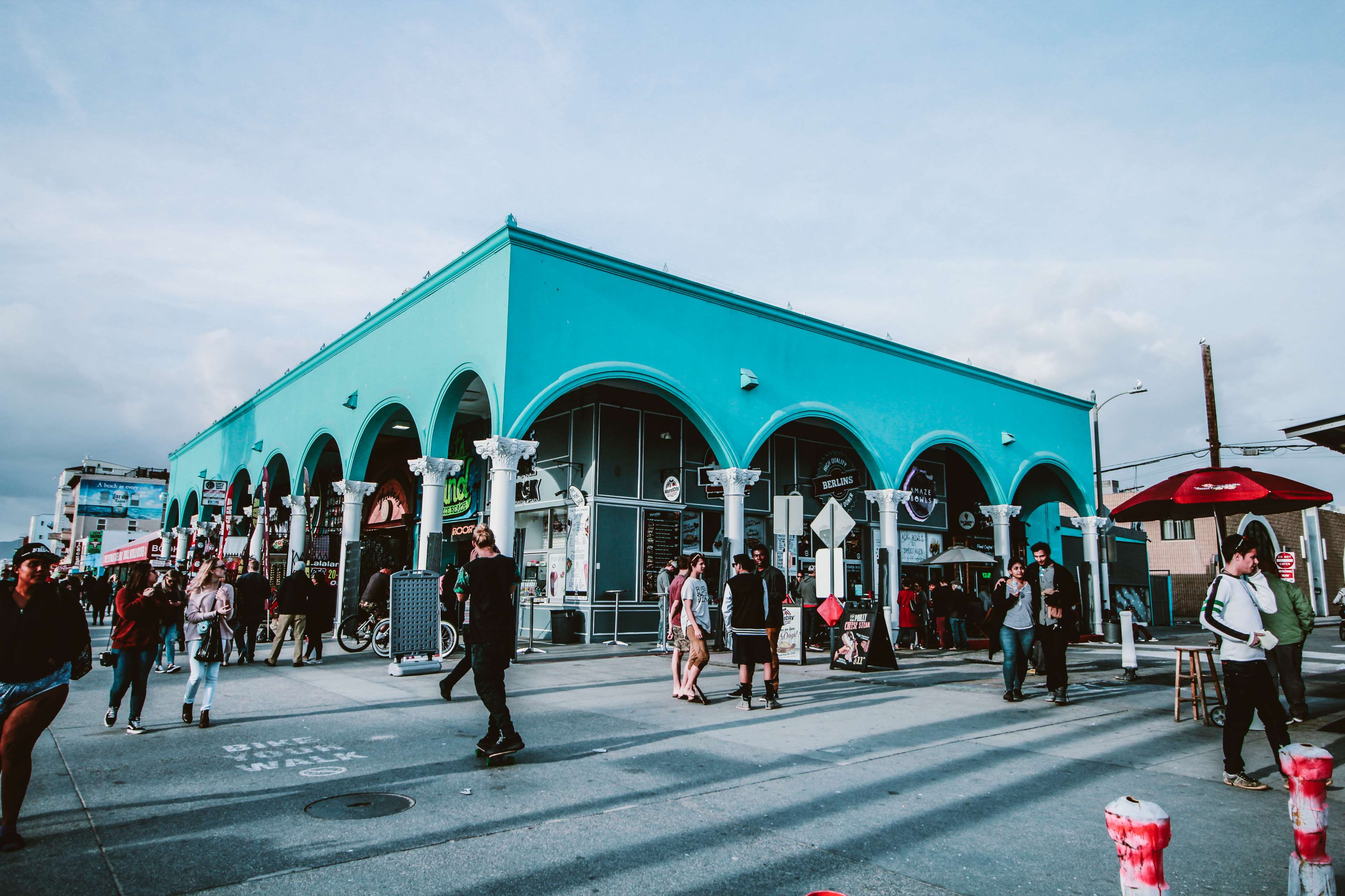 People walking on Venice Beach boardwalk in front of turquoise building 4K HD Wallpaper