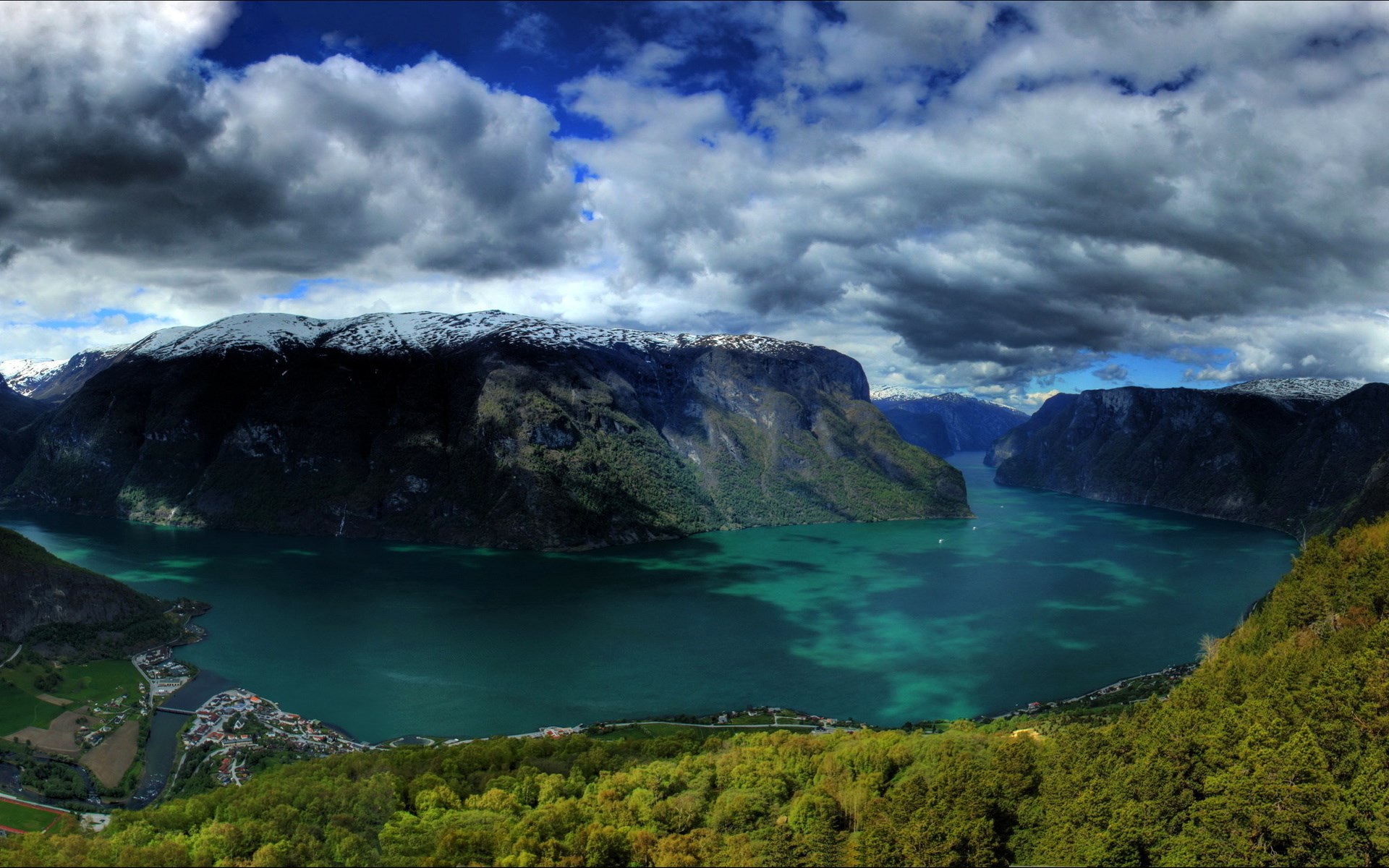 Panoramic view of a fjord with snow-capped mountains and a village HD Wallpaper