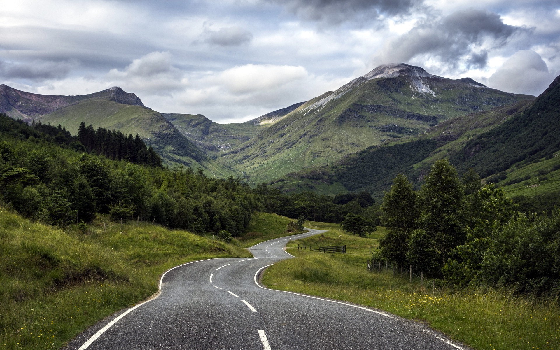 Winding road through a green mountain valley under clouds HD Wallpaper