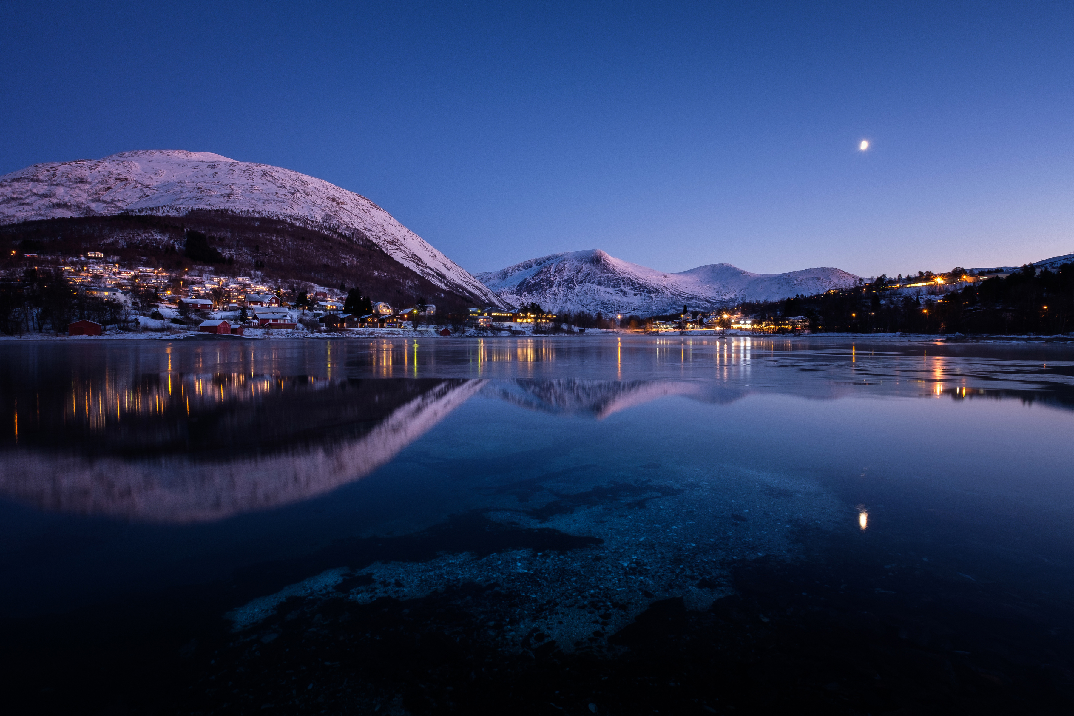 Snowy mountains and city lights reflected in a lake at dusk HD Wallpaper