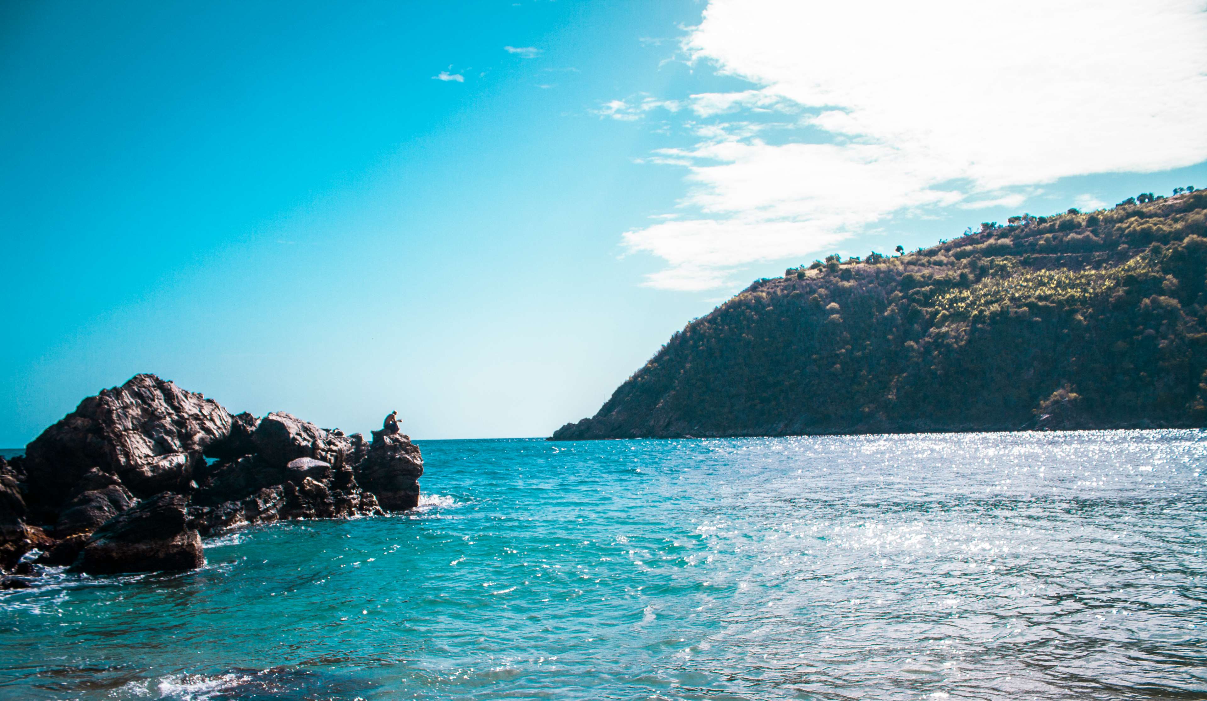 Person sitting on rocks in the ocean near a mountain HD Wallpaper