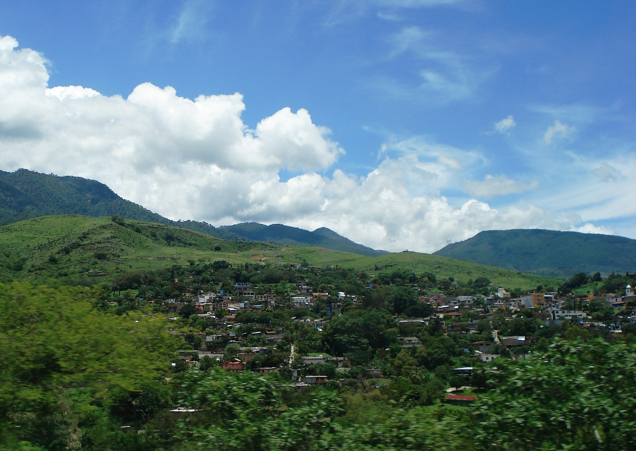 Town nestled in green hills under a blue sky with clouds HD Wallpaper