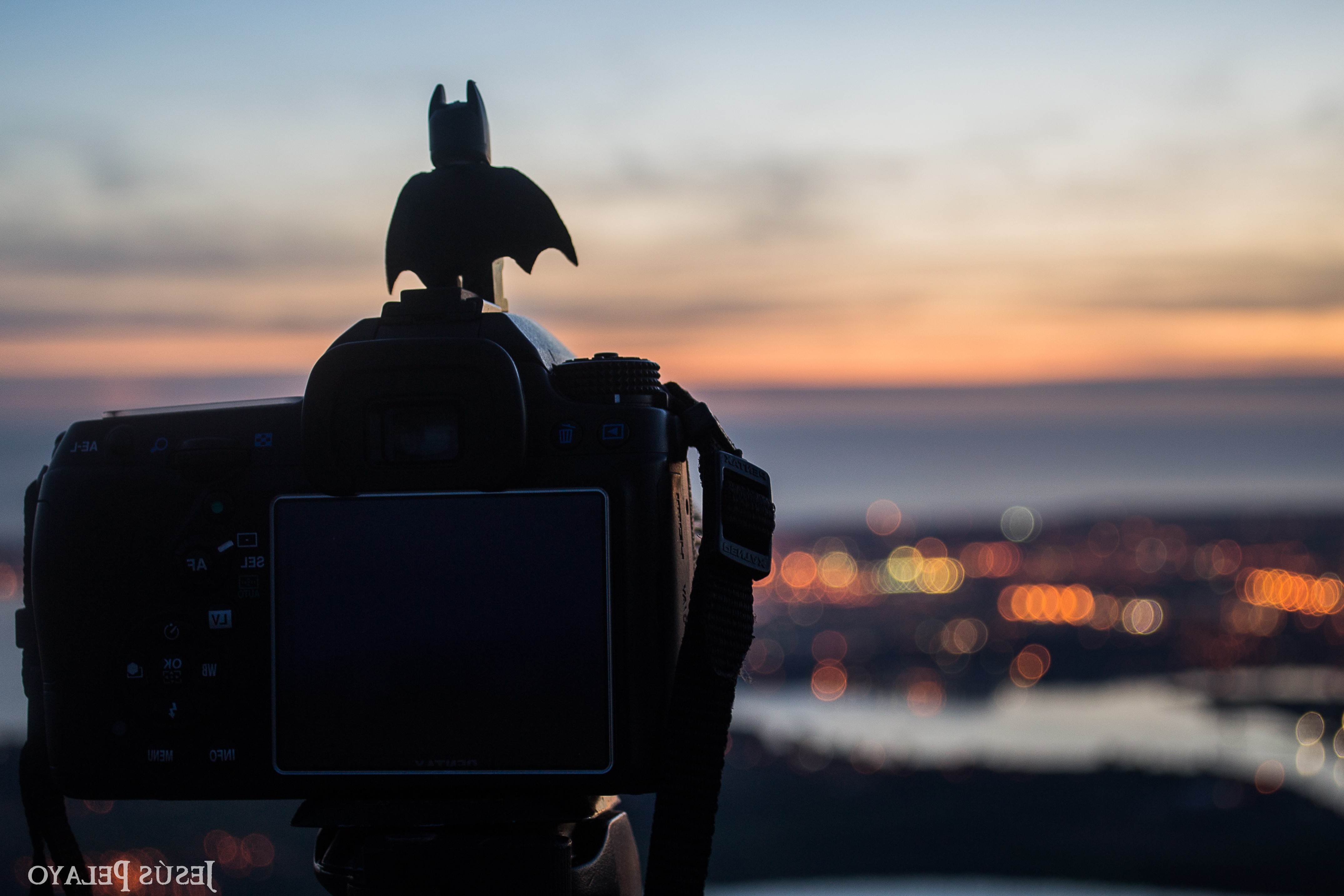 Batman figurine on camera overlooking city at sunset HD Wallpaper