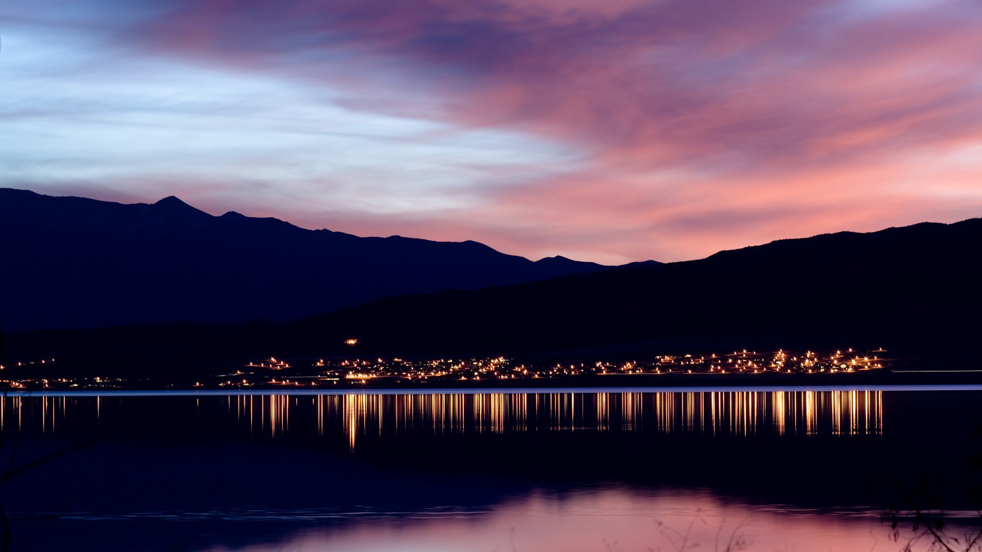 City lights reflected on a calm lake at sunset with mountains HD Wallpaper