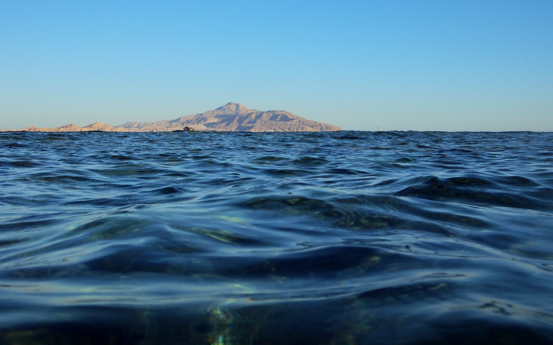 Ocean waves with distant island and mountains under clear sky HD Wallpaper
