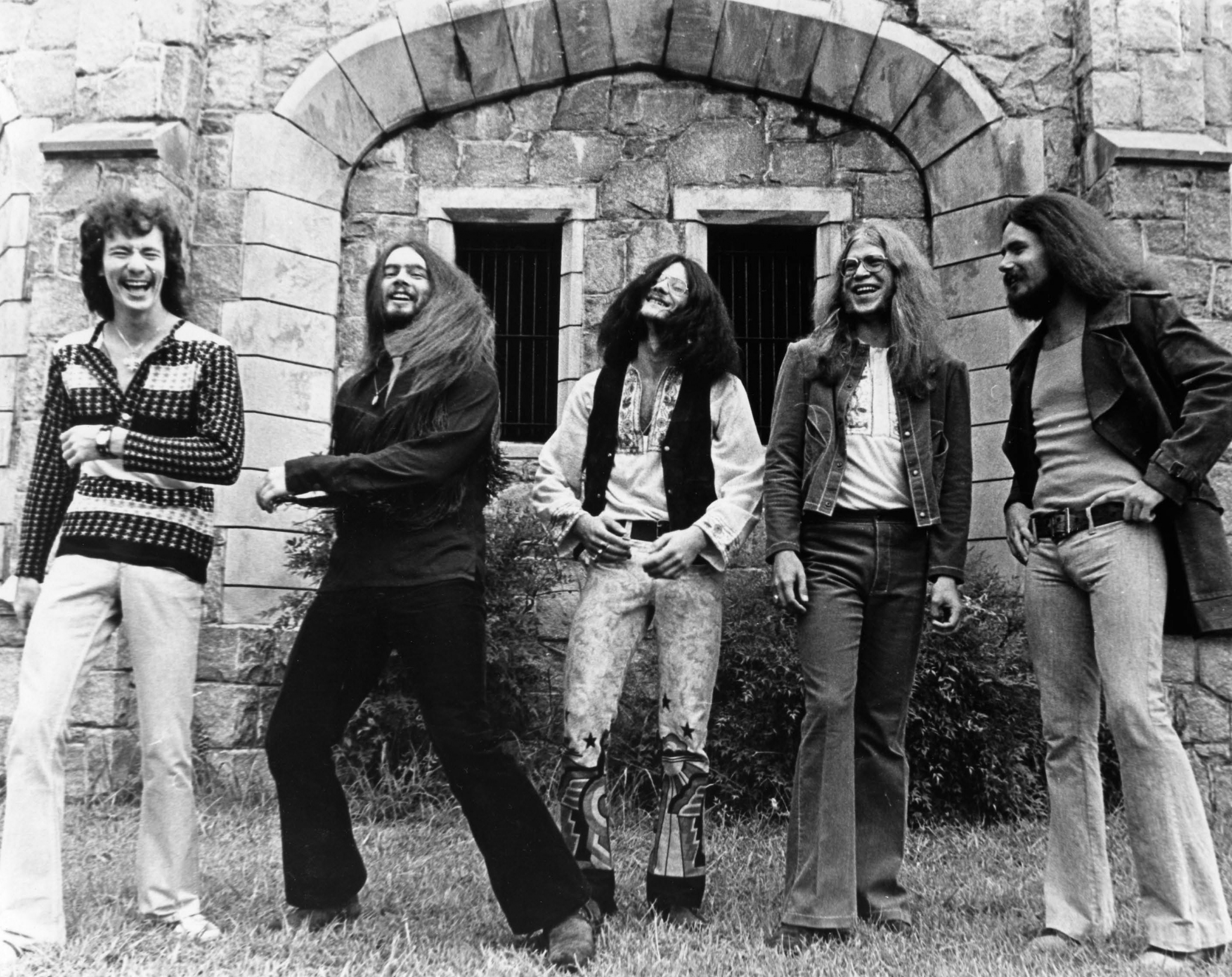 Five men in hippie attire standing in front of a stone building HD Wallpaper