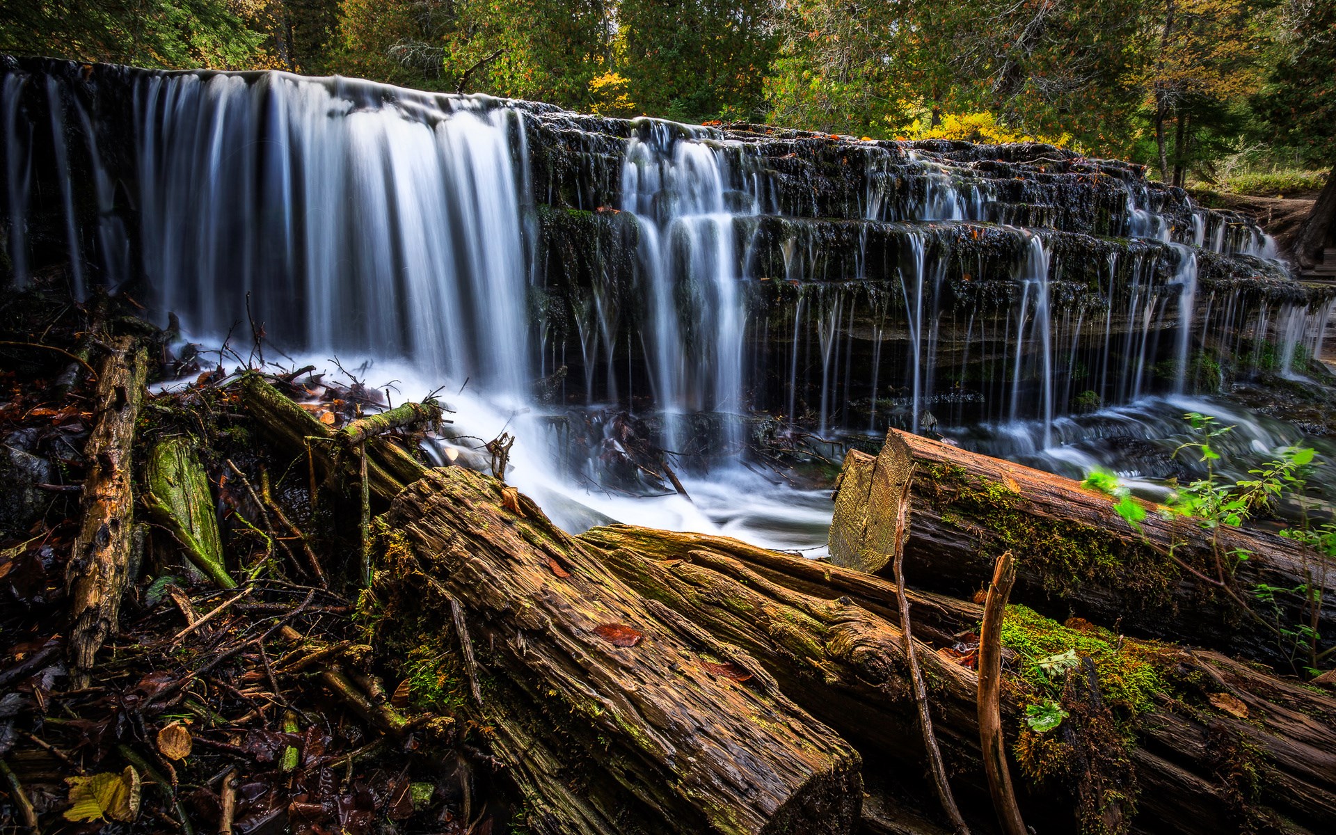 Waterfall cascading over rocks with fallen logs in foreground HD Wallpaper