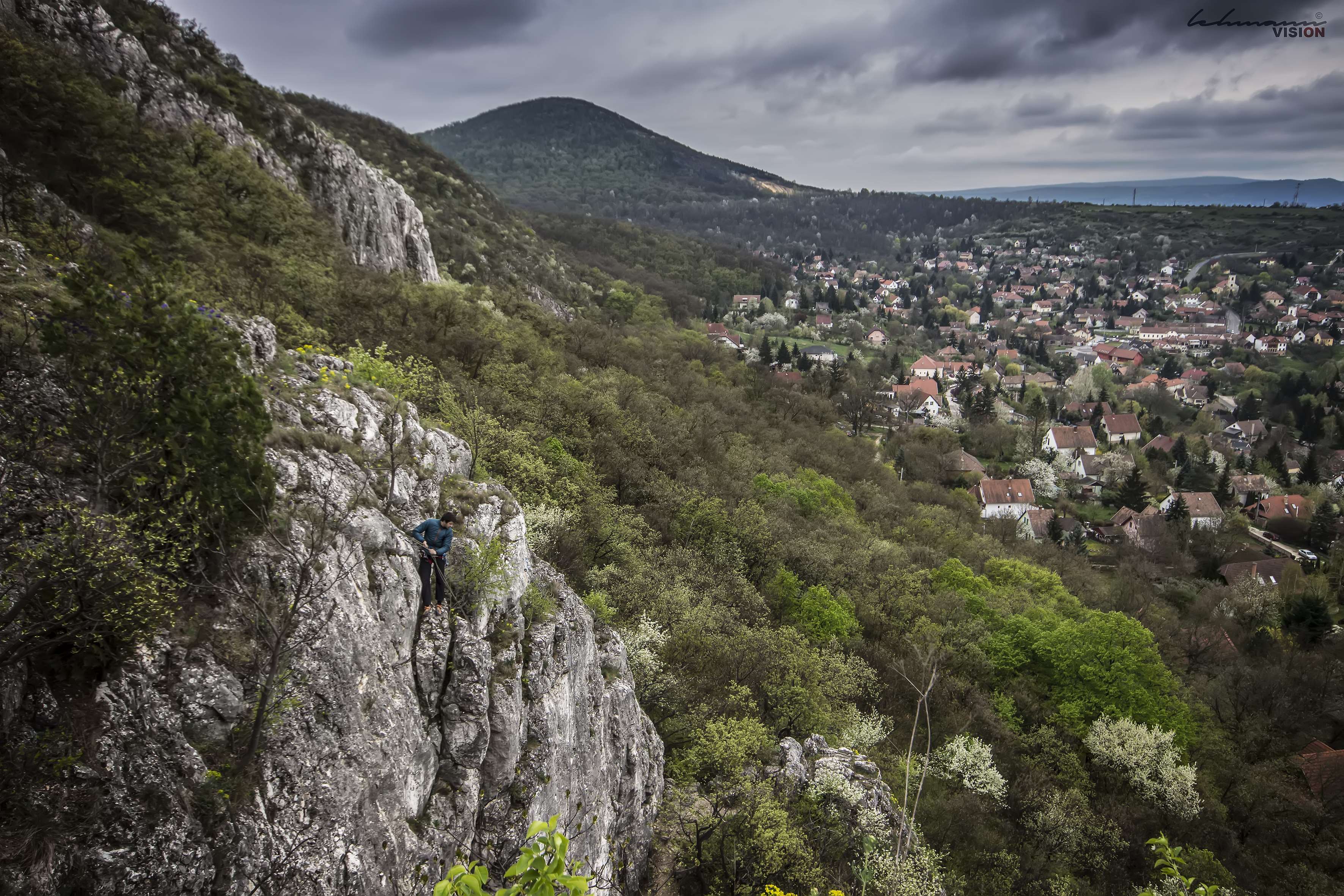 Man climbing on a rocky cliff overlooking a town HD Wallpaper