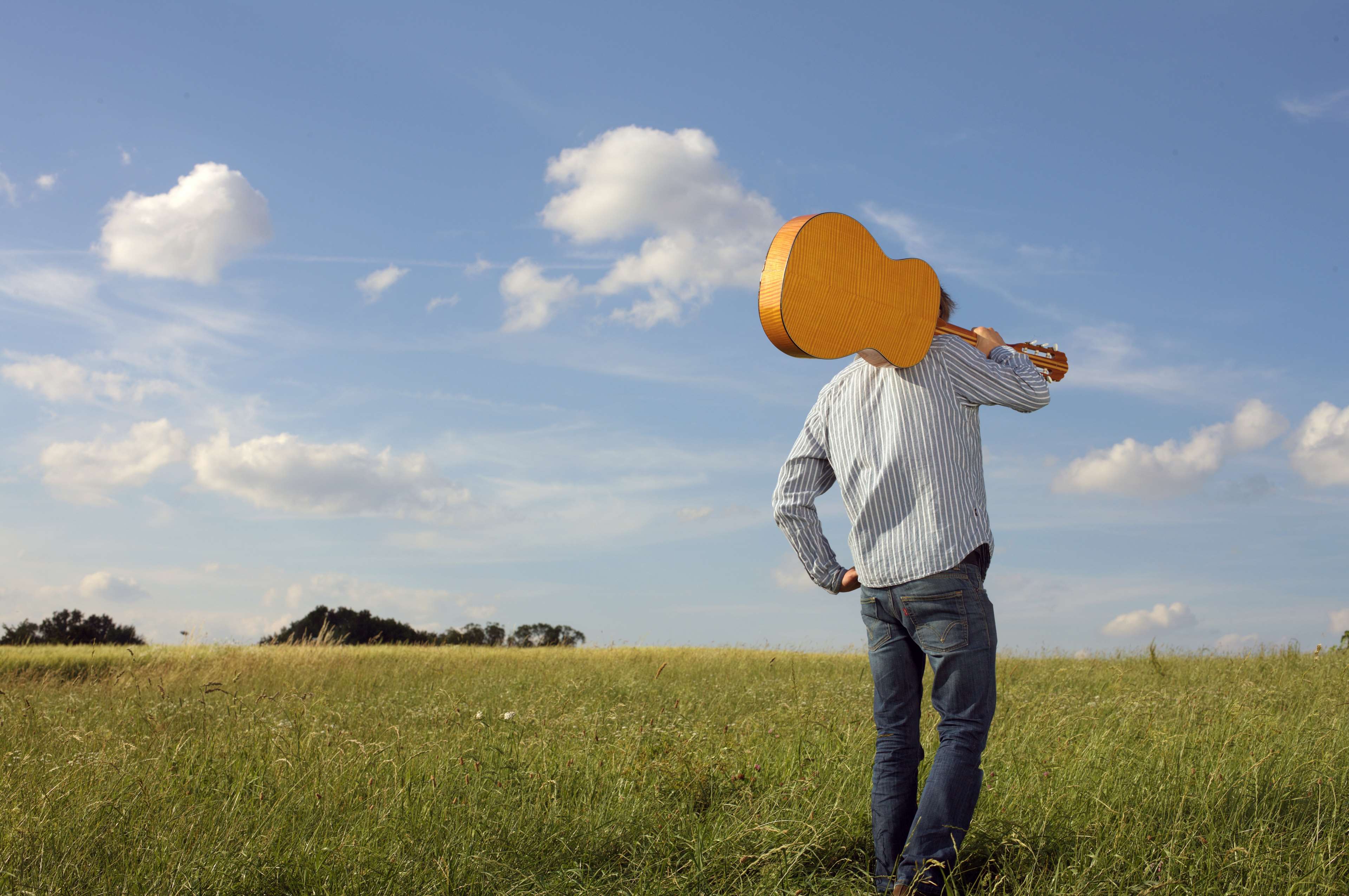 Man carrying a guitar in a grassy field under a blue sky HD Wallpaper