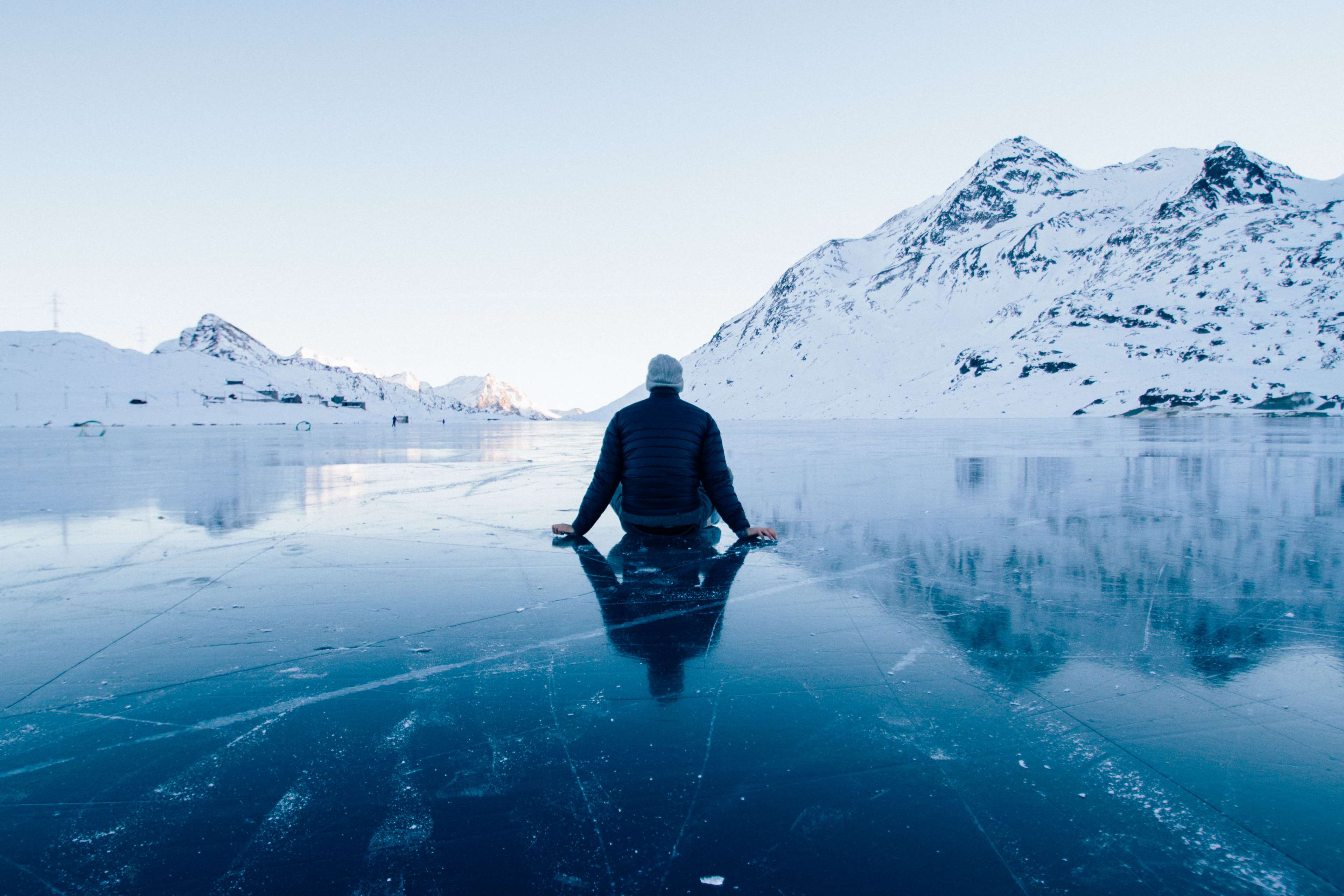 Man sitting on a frozen lake with snow-capped mountains HD Wallpaper