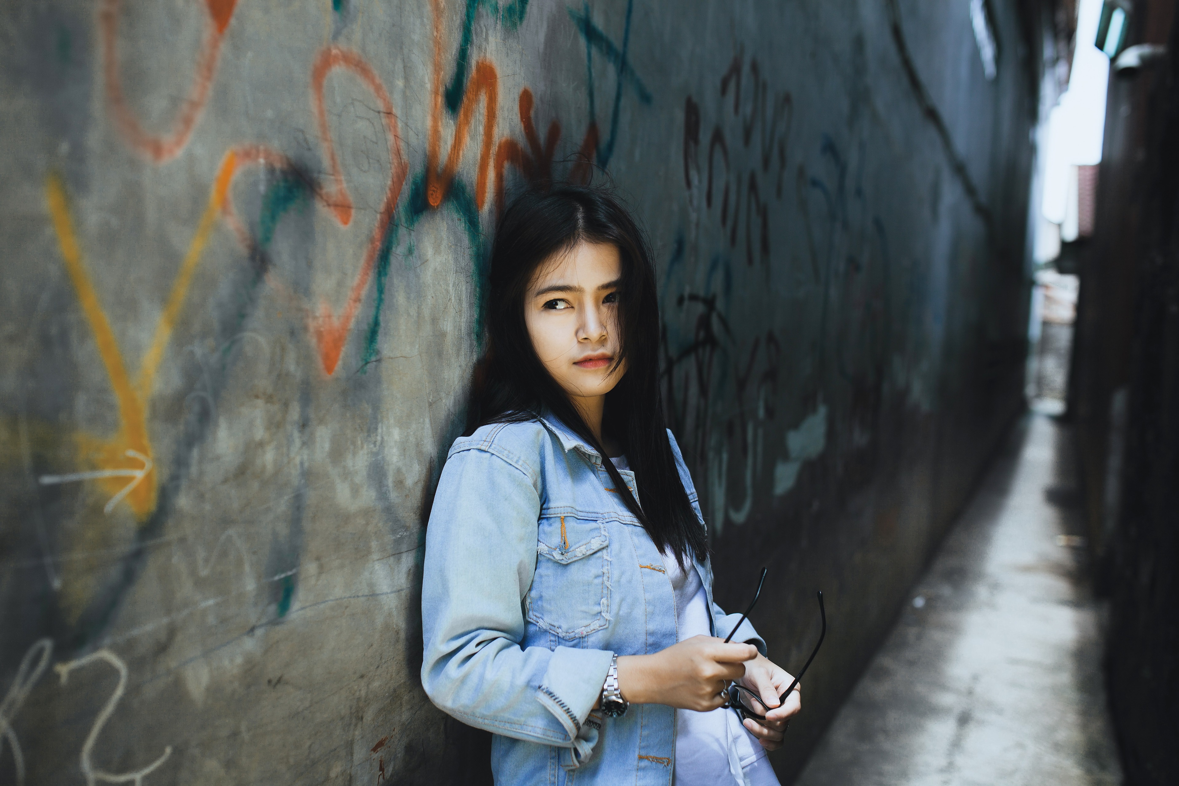 Asian girl in denim jacket leaning against graffiti wall HD Wallpaper