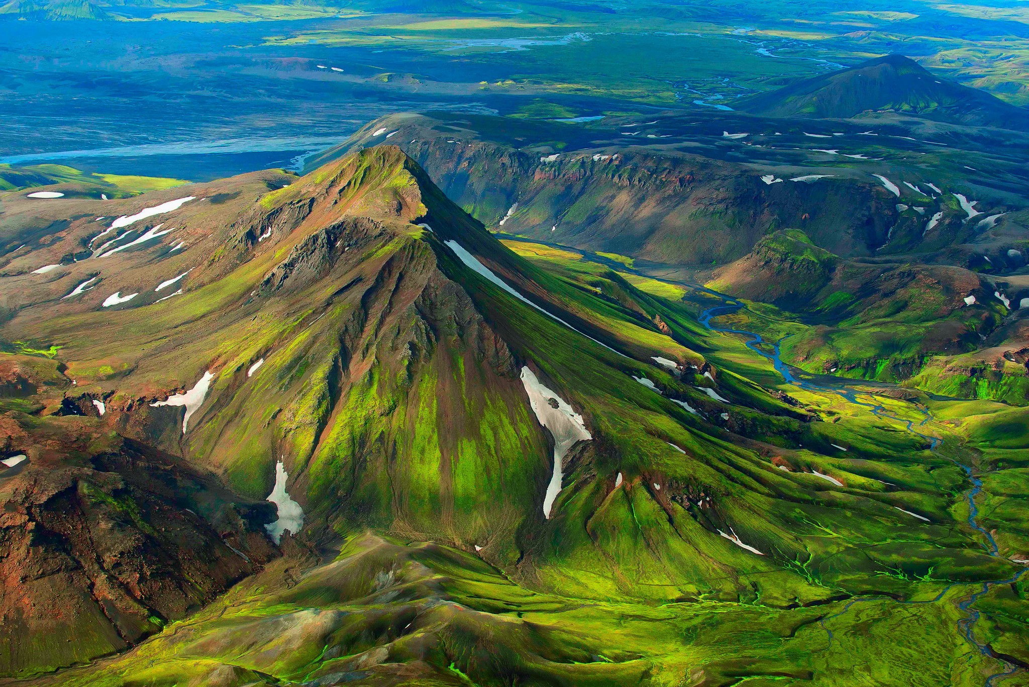 Aerial view of a green mountain range with snow patches and a river HD Wallpaper