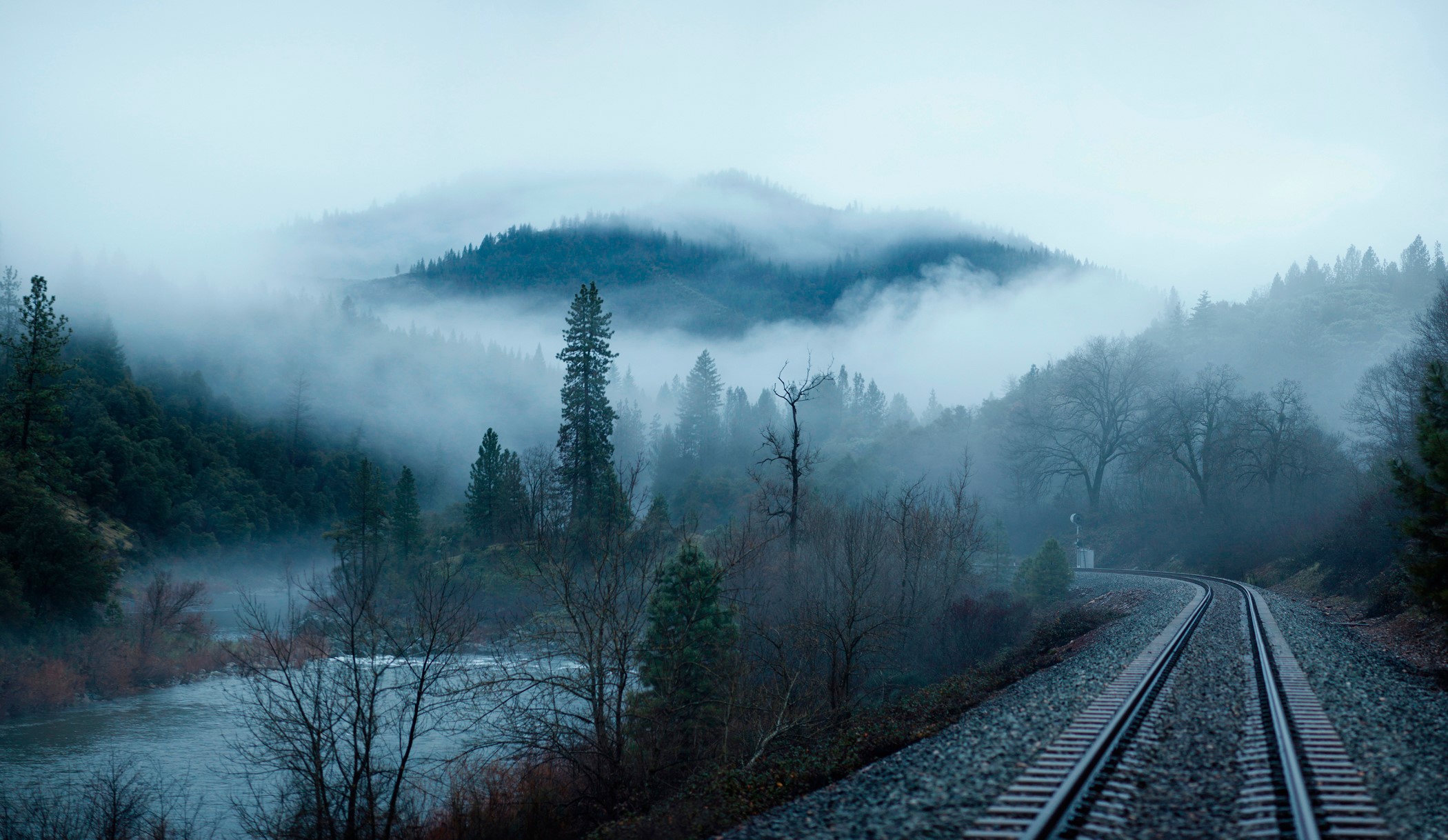 Train tracks curve through a foggy forest with a river HD Wallpaper