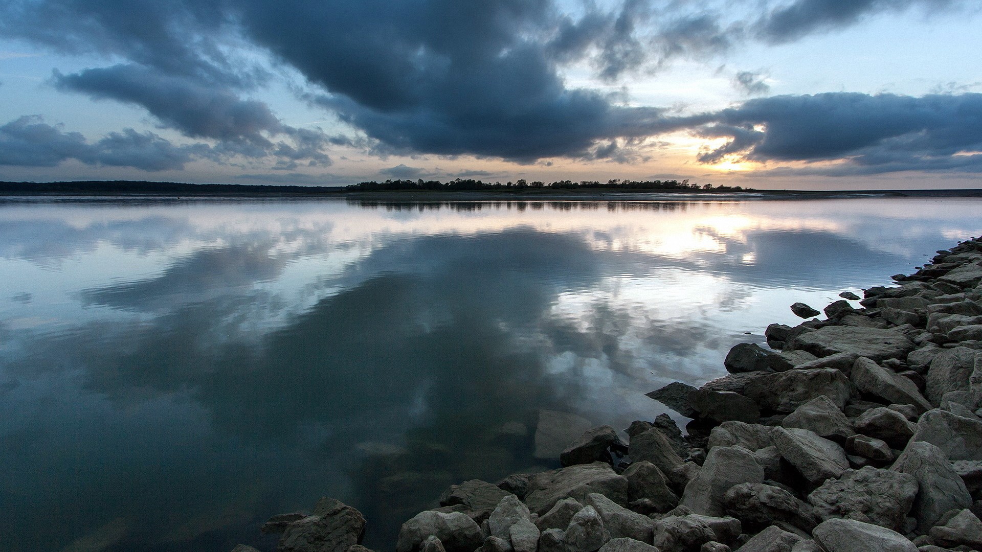 Calm lake with rocky shore and cloudy sky at sunset HD Wallpaper
