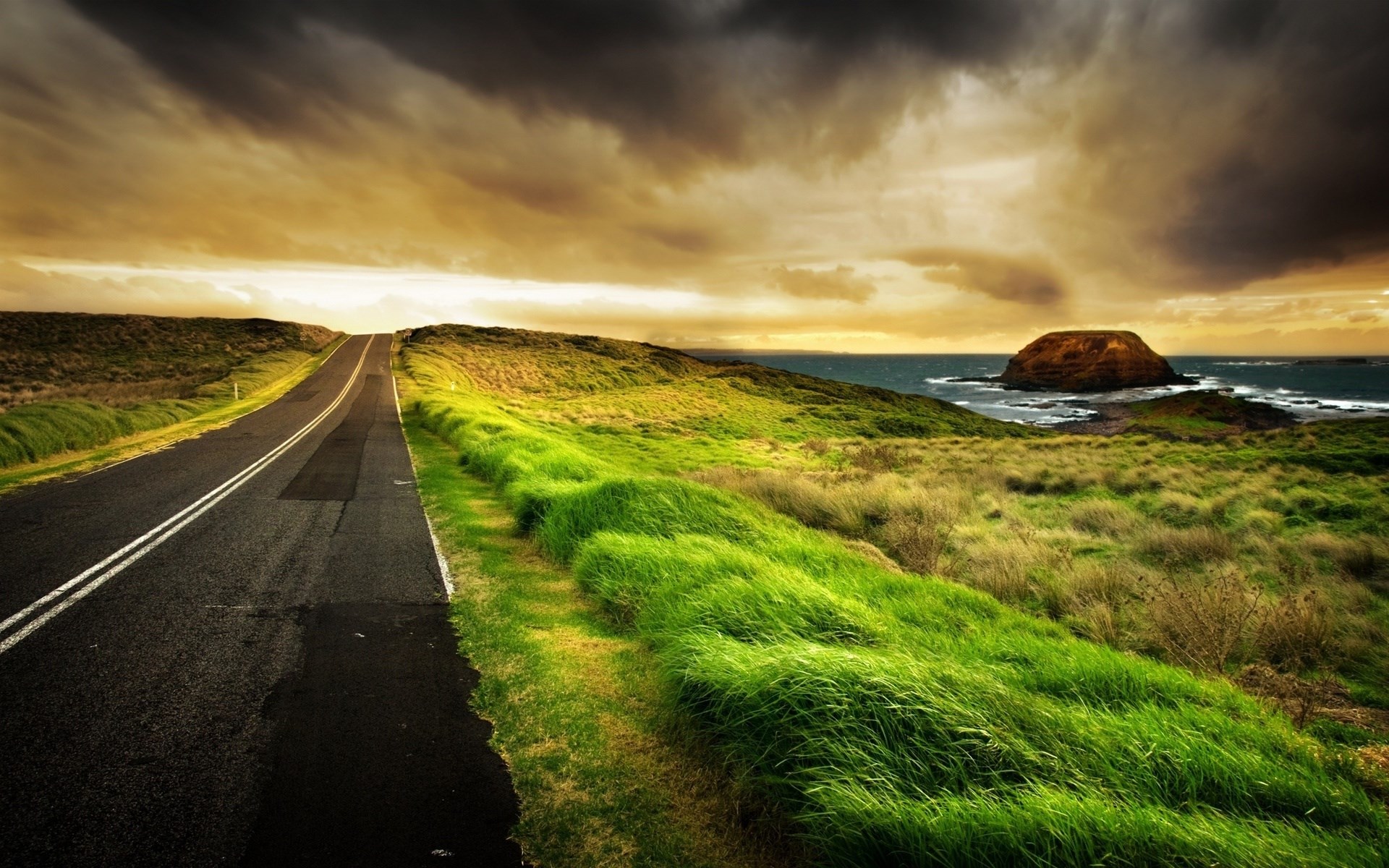 Road leading to the ocean with a rock formation HD Wallpaper