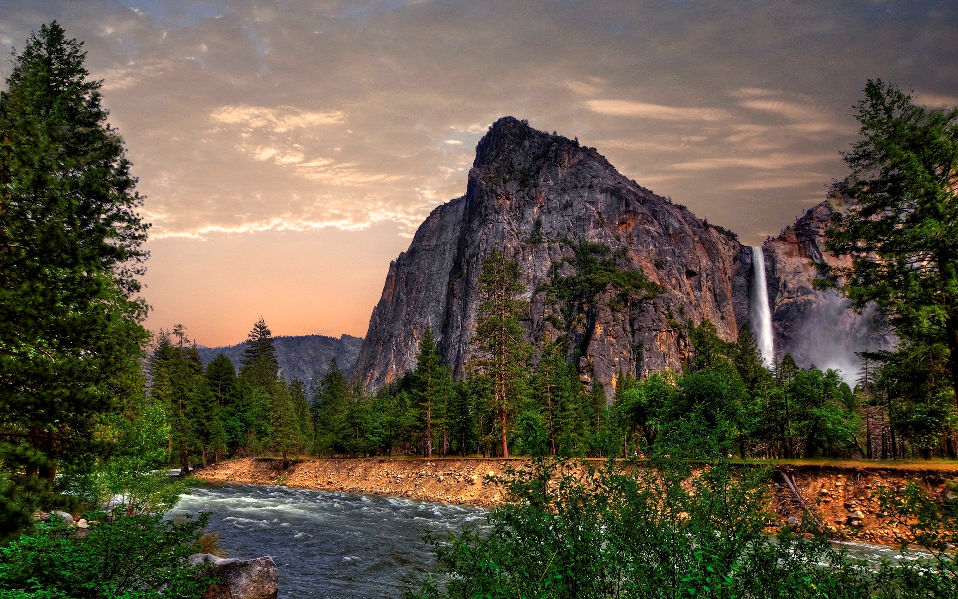 Waterfall cascading down a rocky cliff face near a river and pine trees HD Wallpaper