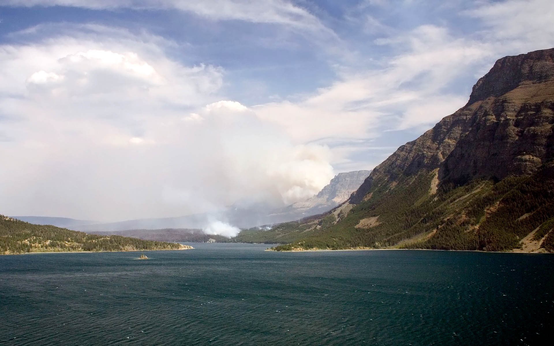 Smoke rising from a wildfire over a lake and mountains HD Wallpaper