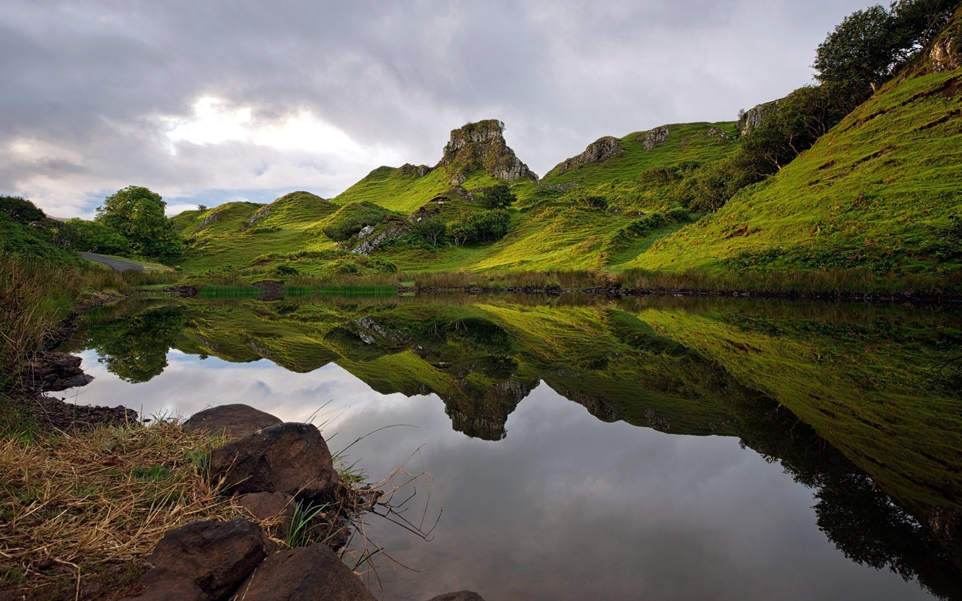 Green hills reflected in a calm lake under a cloudy sky HD Wallpaper