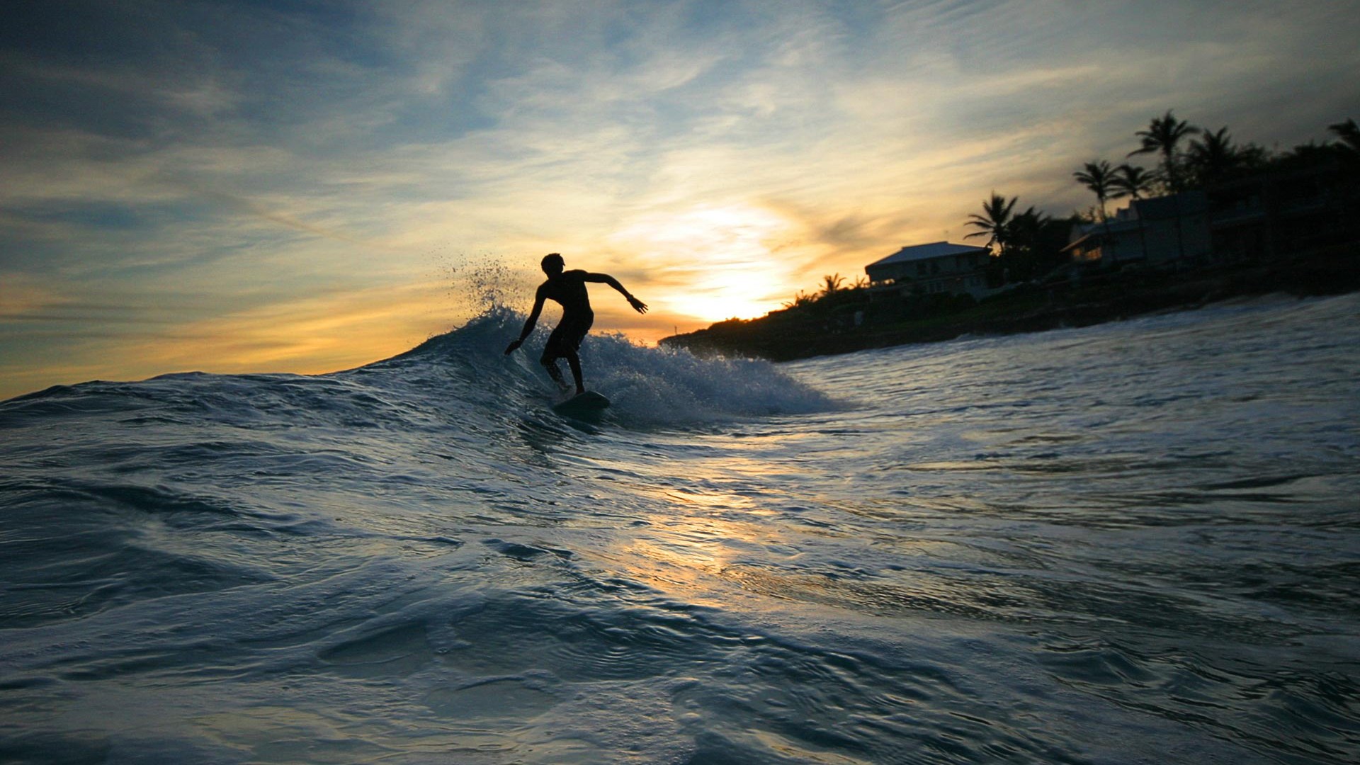 Surfer riding a wave at sunset with houses on a hill HD Wallpaper