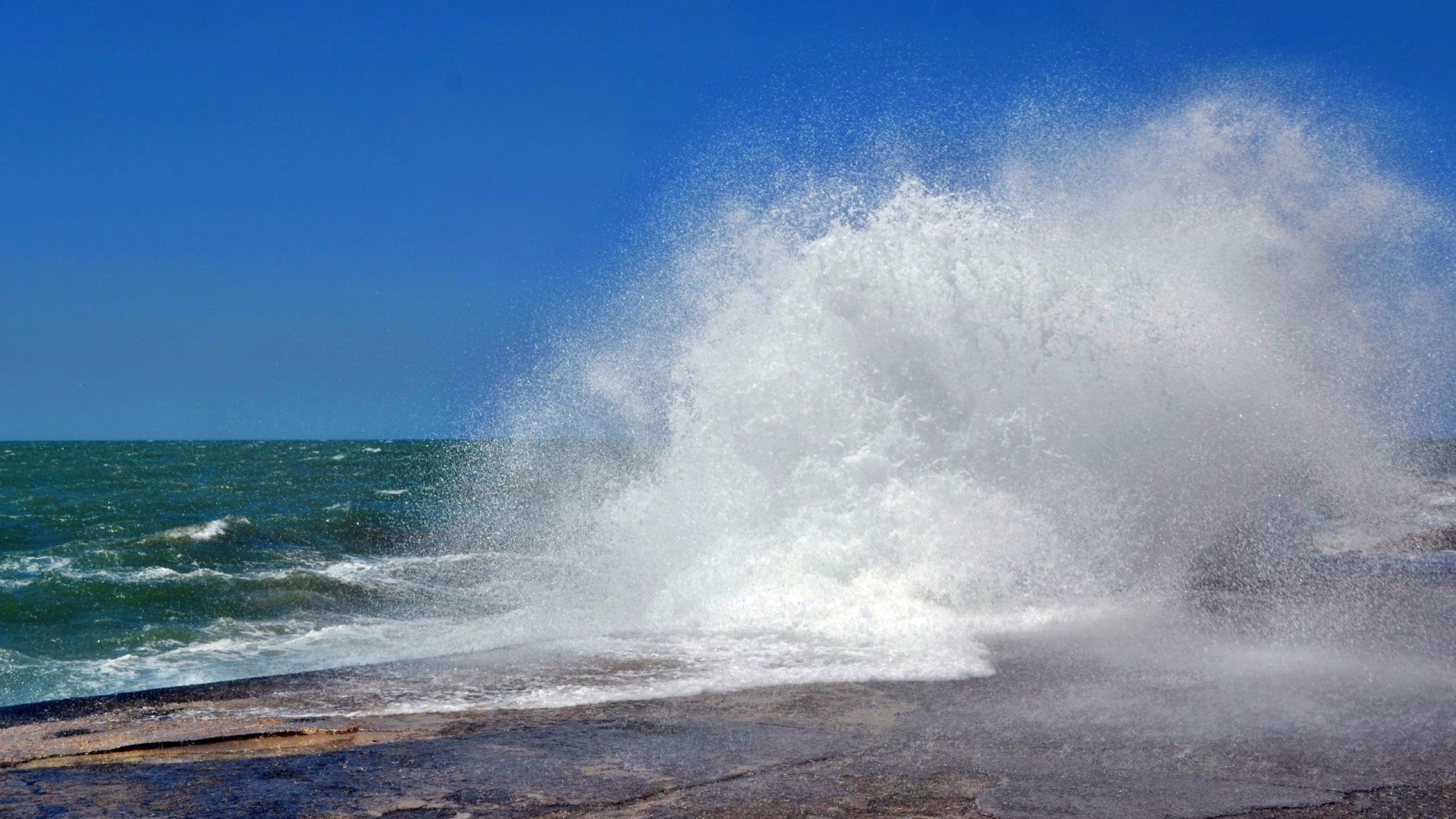Wave crashing on a rocky shore under a blue sky HD Wallpaper