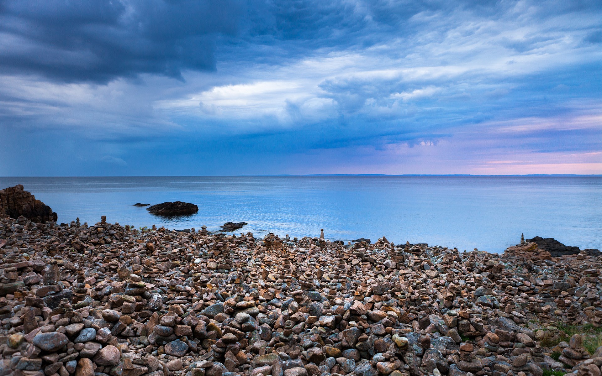Stone cairns on a rocky shore with a calm sea and cloudy sky HD Wallpaper