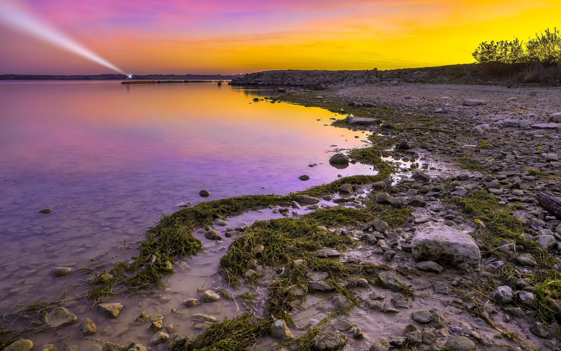 Sunset over a rocky shore with a lighthouse beam HD Wallpaper