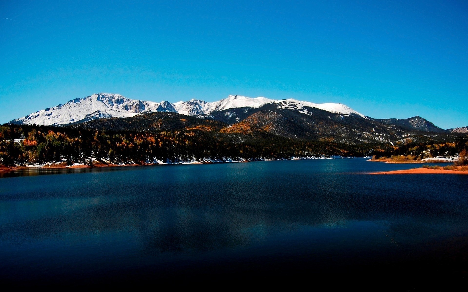 Snow-capped mountains overlook a dark blue lake and autumn forest HD Wallpaper