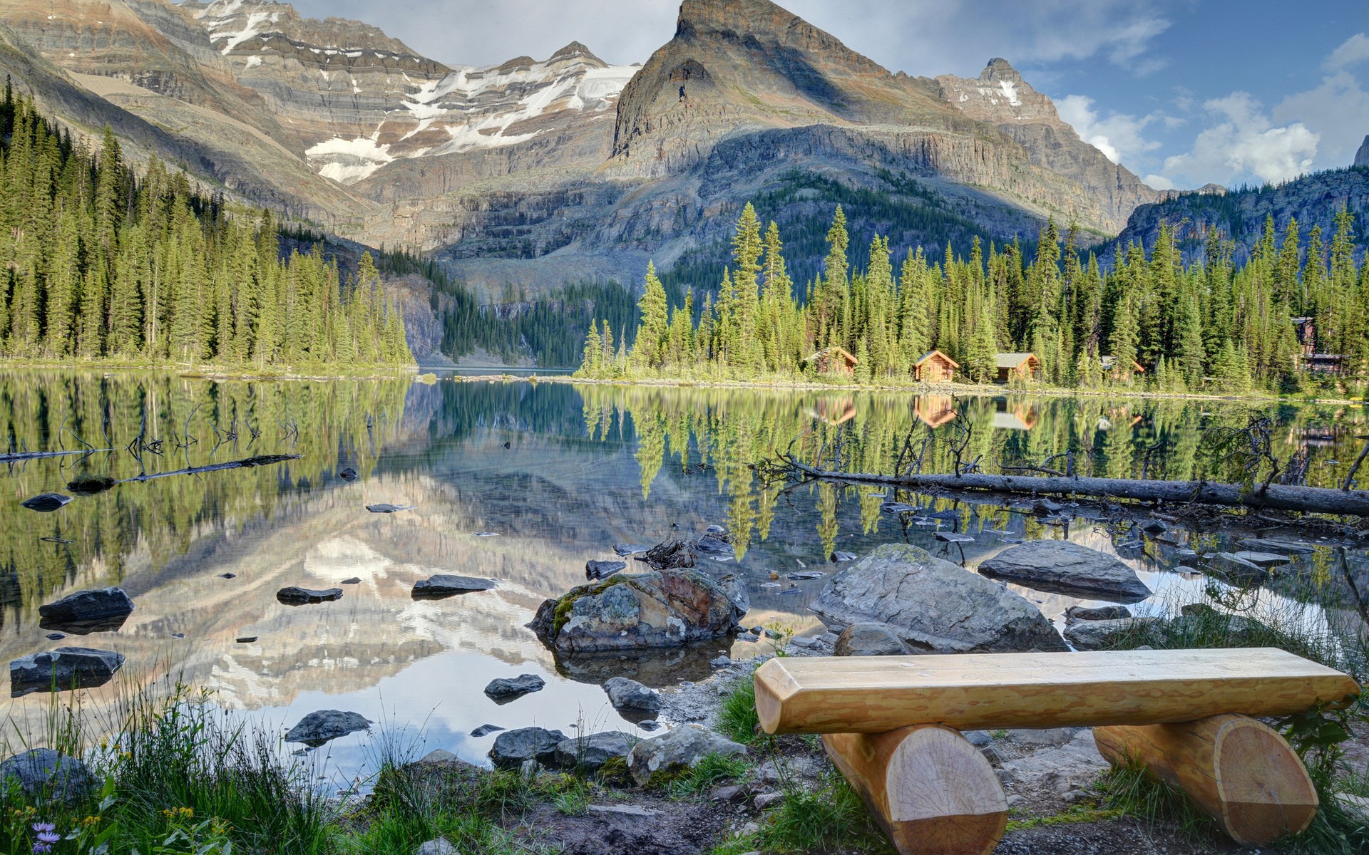 Wooden bench by a calm lake with mountains and cabins HD Wallpaper